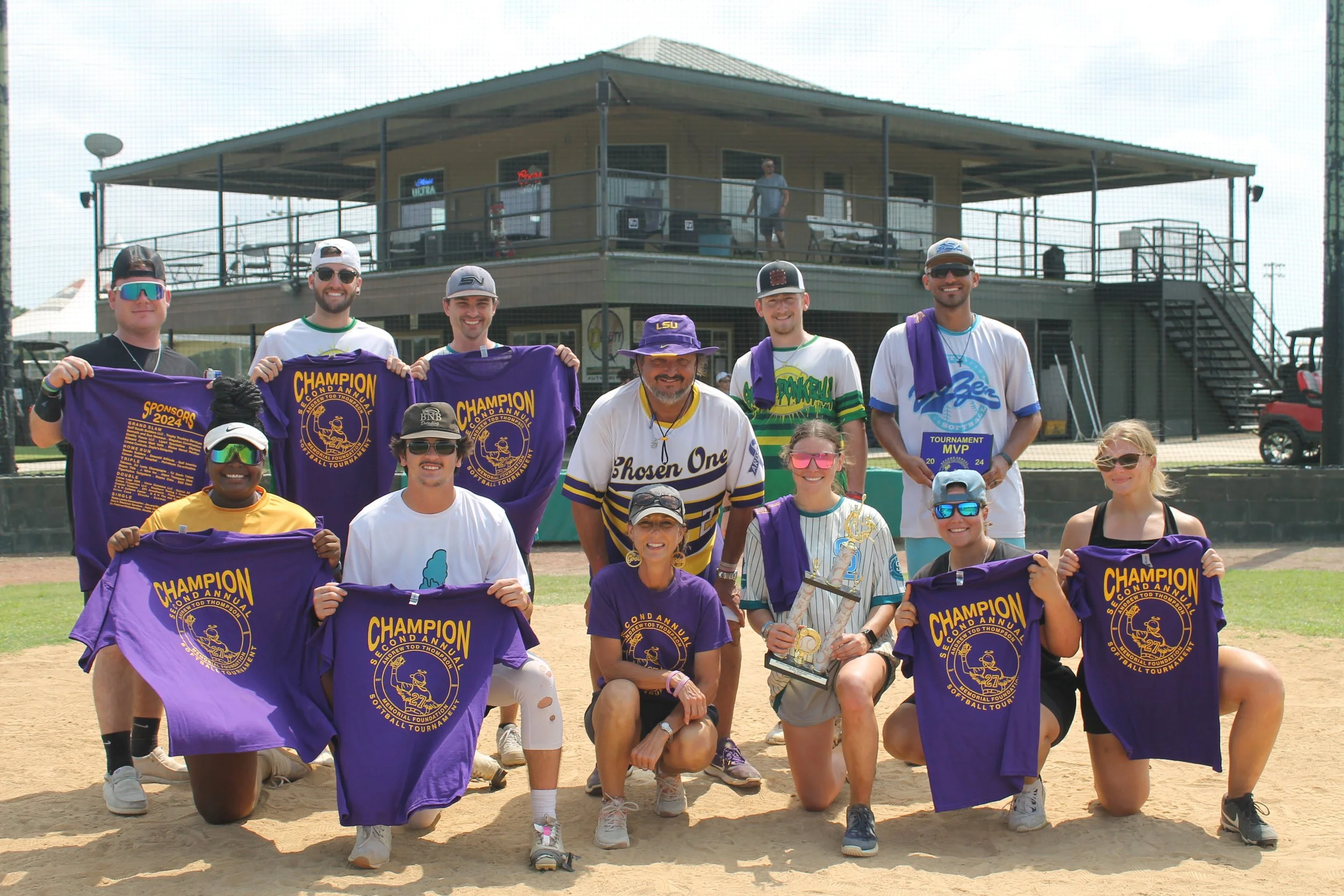 Group of people on a baseball field celebrating a victory, holding purple champion and tournament T-shirts and trophies, with a clubhouse building in the background.