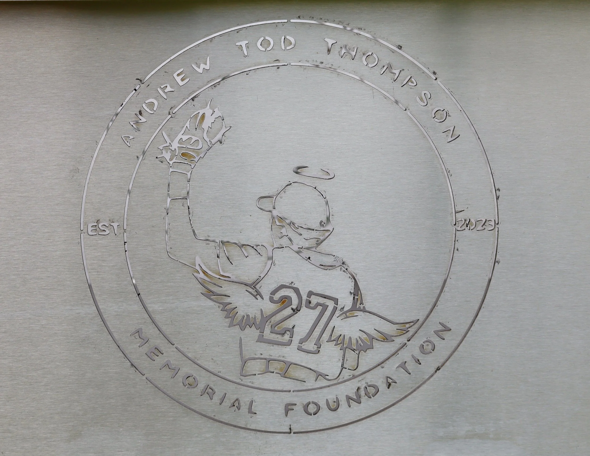 A metallic surface with an engraved logo of a bear holding a fish, surrounded by circular text. The text reads: 'Andrew Todd Two Ton Son', 'Est 2023', 'The Original Foundation', and the number '27' at the bottom.