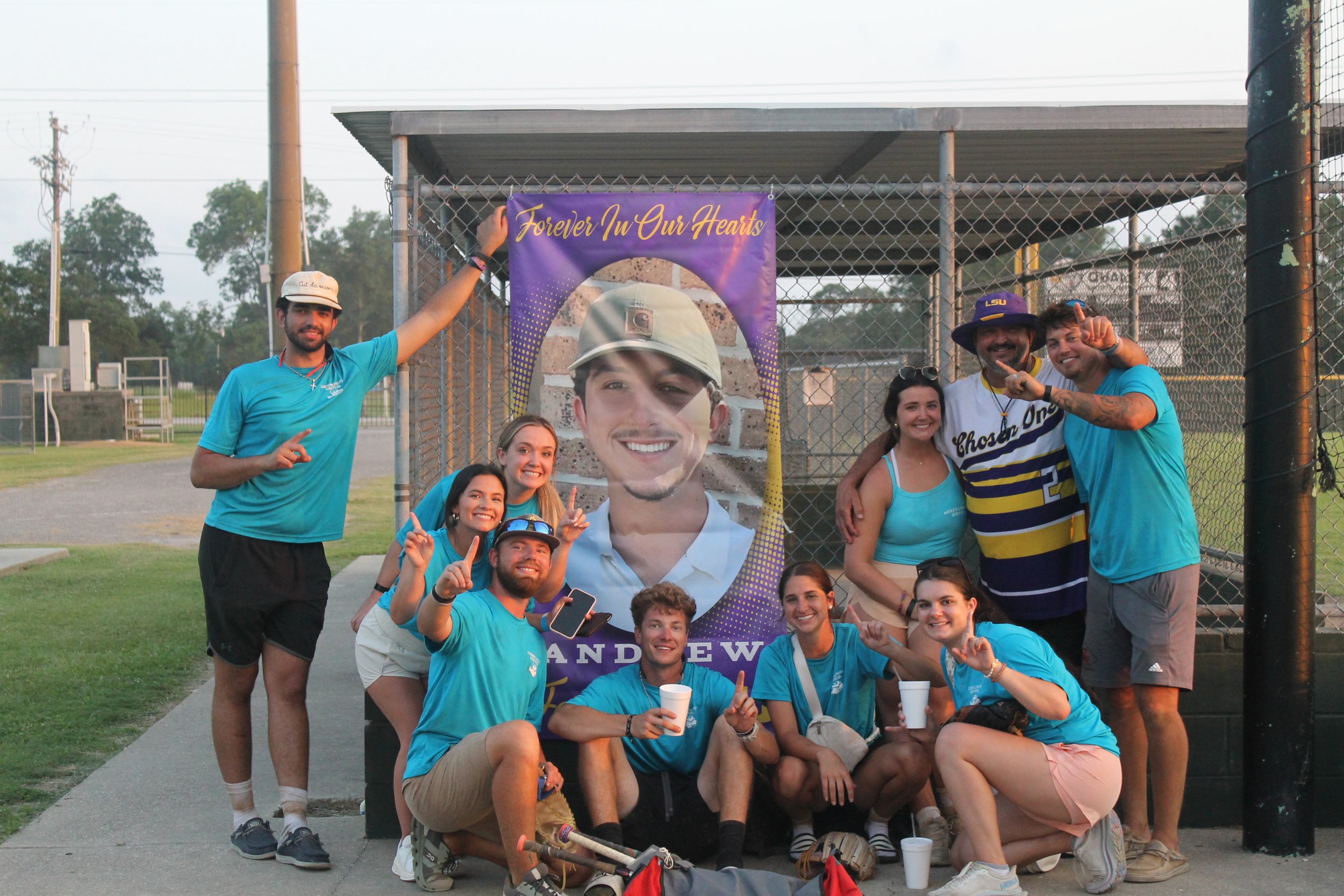 Group of people posing in front of a large poster with a smiling man in a helmet, with the text "Forever In Our Hearts" and "Andrew". The group is making peace signs and smiling, some wearing matching blue shirts.