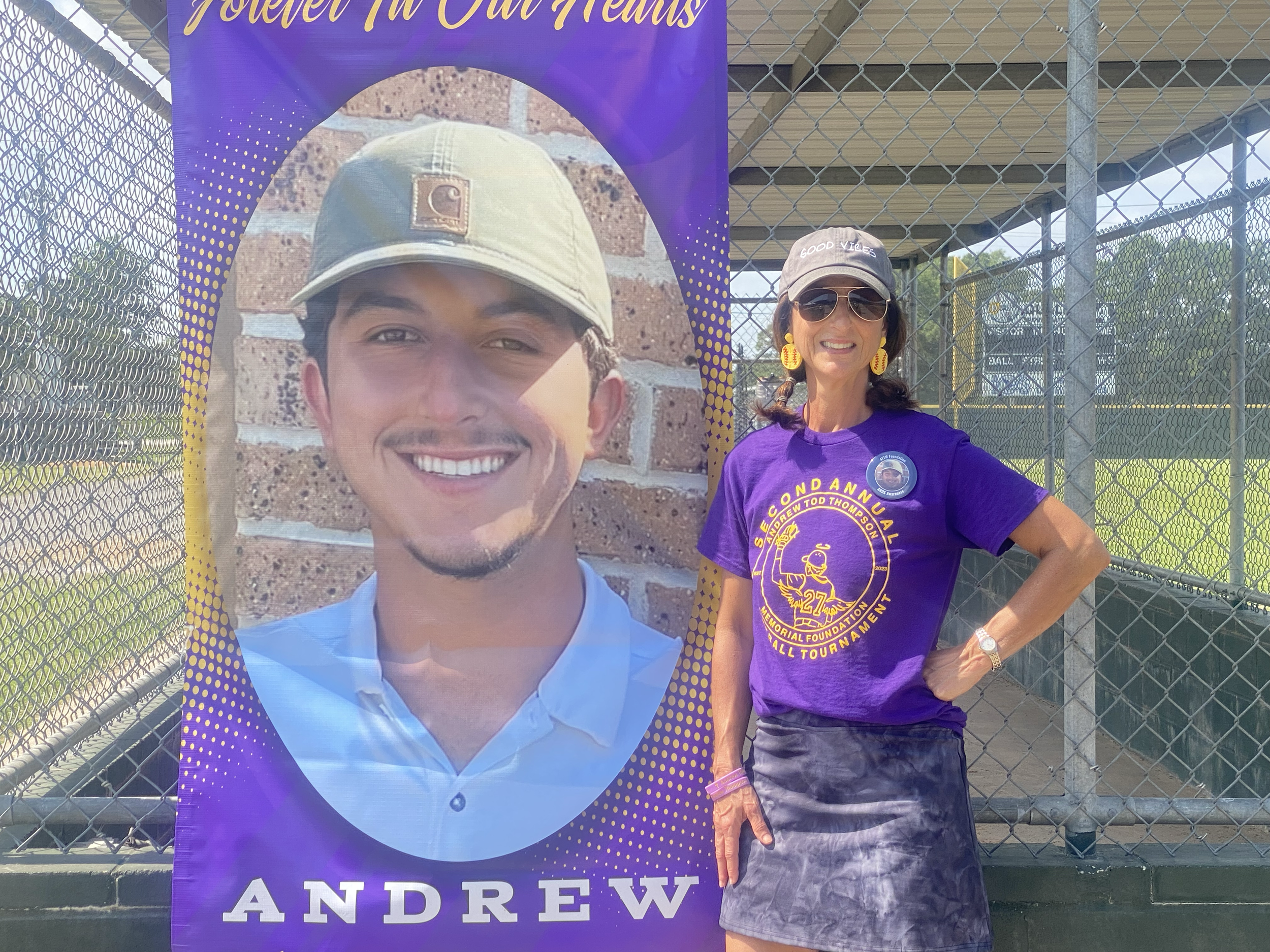 A woman wearing sunglasses, a gray cap with 'Good Vibes' written on it, a purple T-shirt with yellow text and graphics, a black patterned skirt, and yellow earrings, standing next to a large poster of a smiling young man in a baseball cap. The woman 