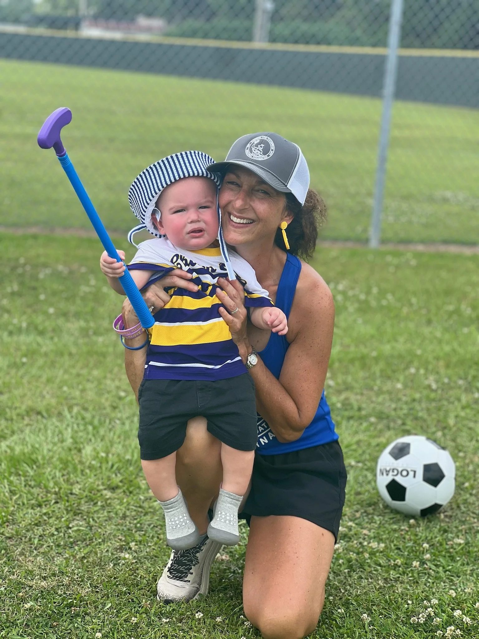 A woman kneeling on grass holding a young boy, both smiling. The boy is holding a purple and blue golf club, wearing a striped navy and yellow shirt, black shorts, and gray socks. The woman is wearing a blue tank top, black shorts, a gray cap, and ye