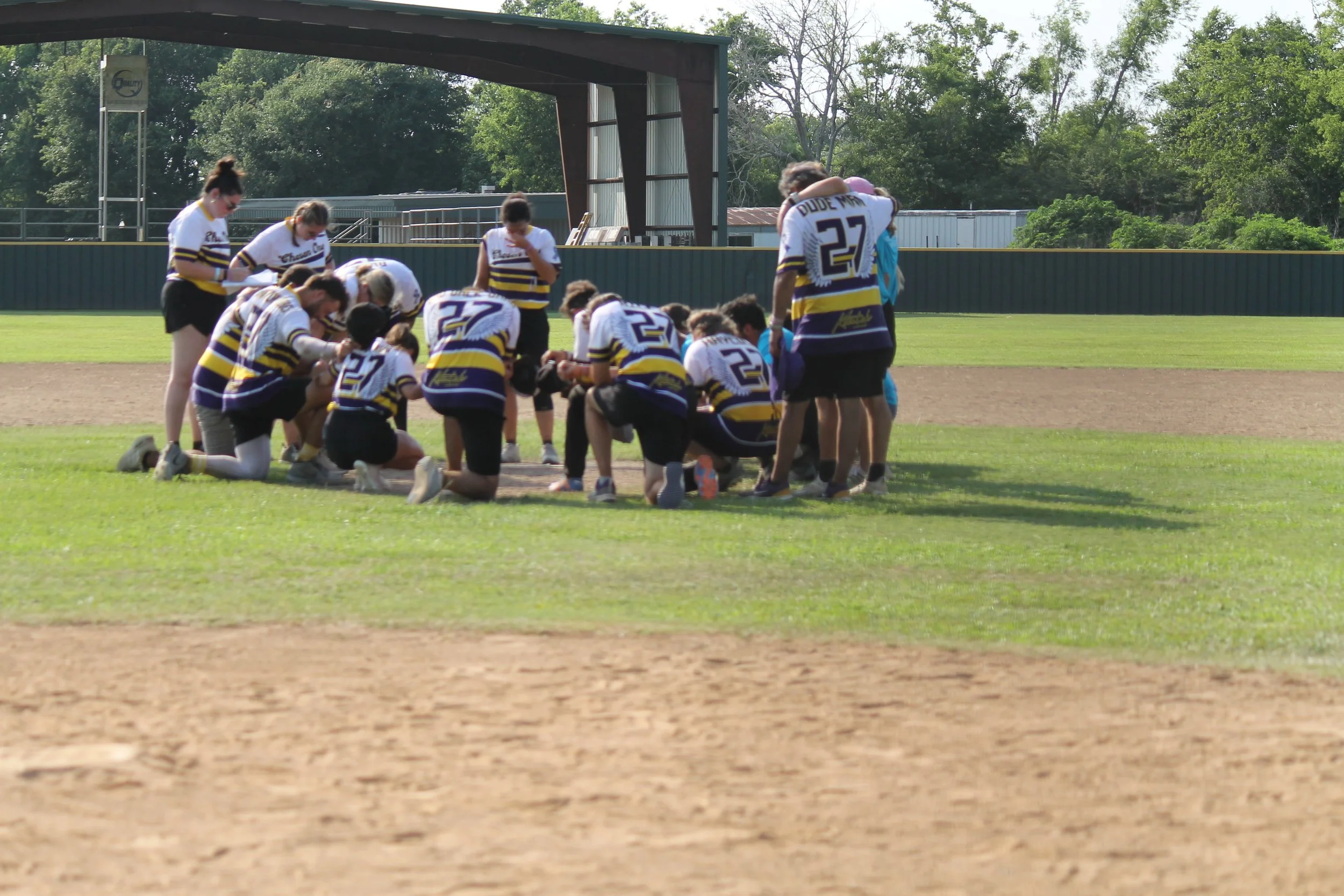 A group of girls and women on a baseball field, gathered in a prayer circle or team huddle, wearing baseball uniforms with purple, yellow, and white colors, on a sunny day.
