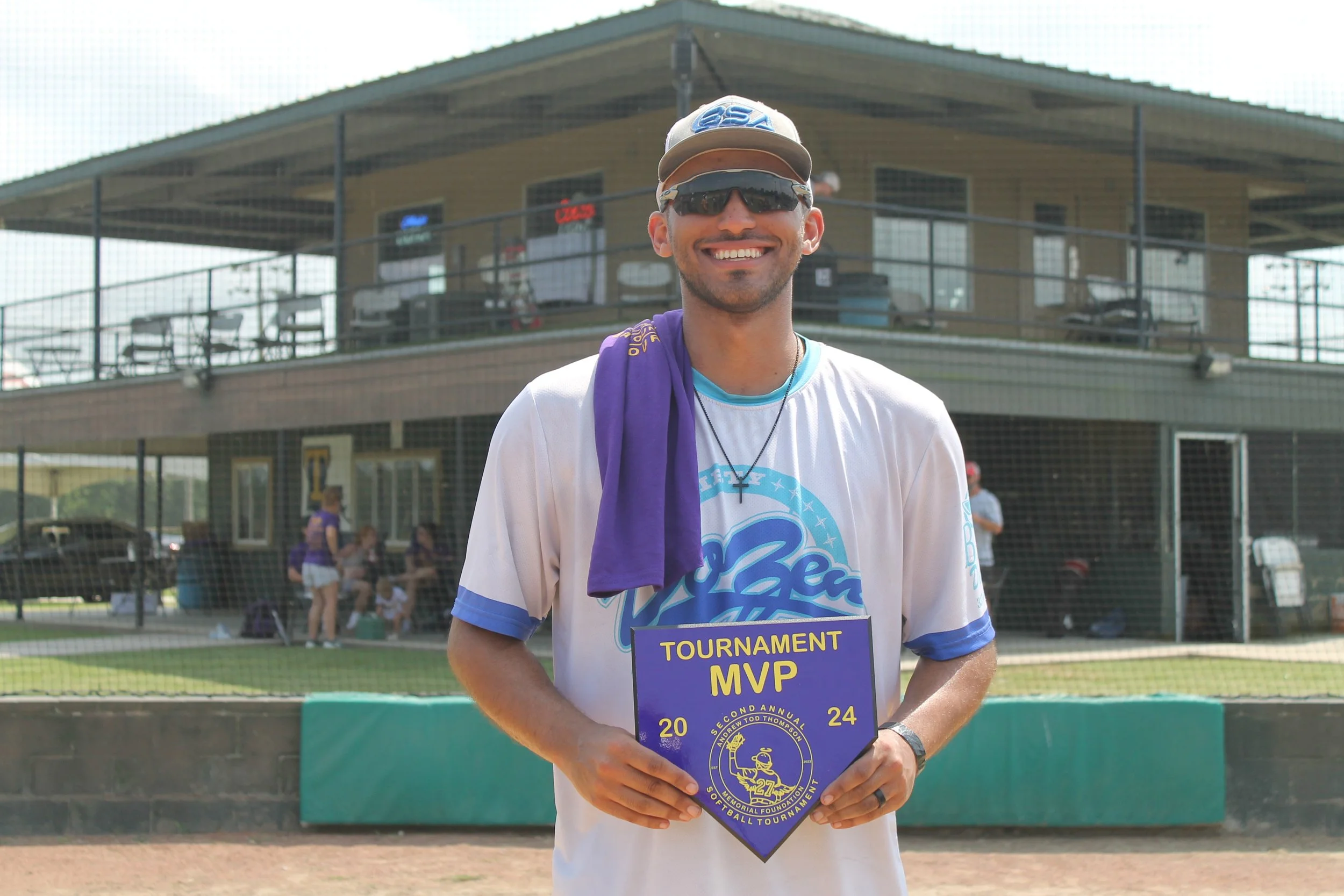 Smiling man wearing sunglasses and a baseball cap, holding a purple MVP award plaque, standing on a baseball field with people and a building in the background.