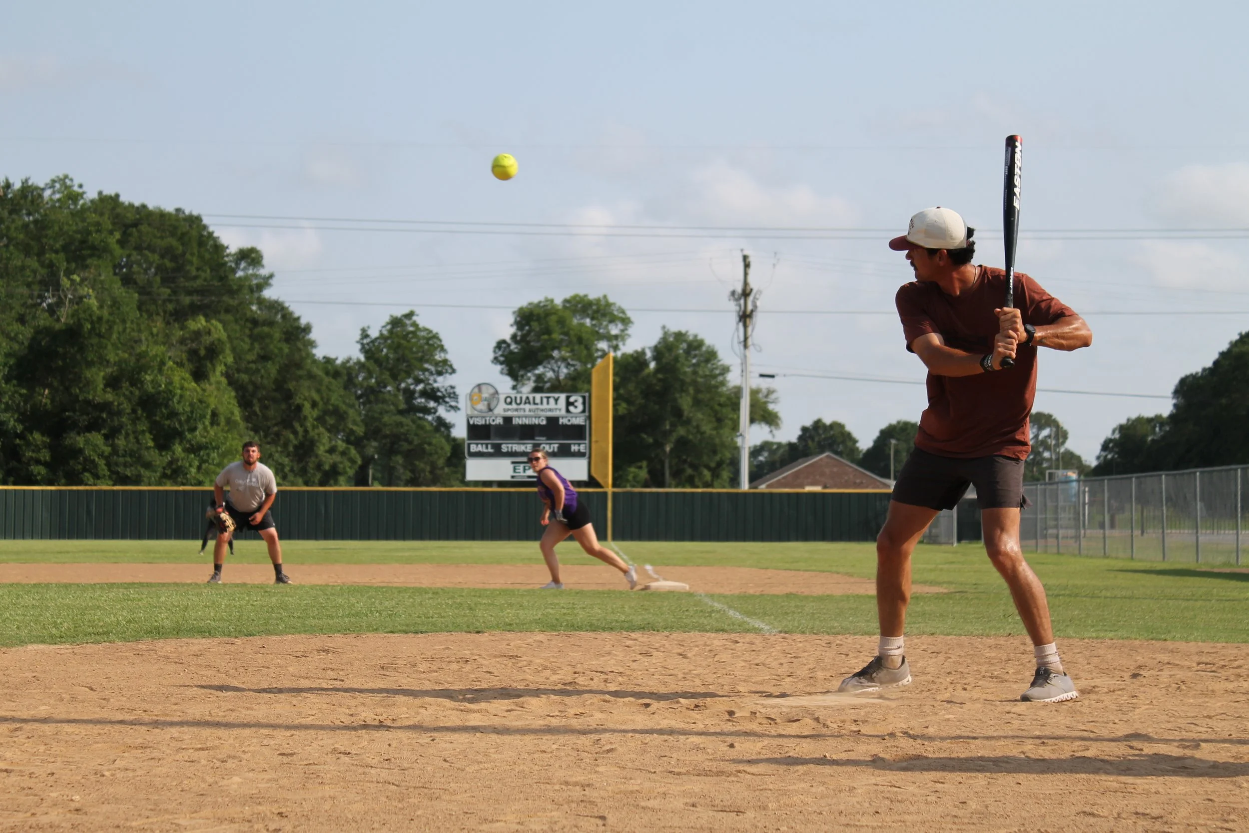 A group of people playing baseball on a field during the daytime with the batter preparing to hit the ball, the pitcher ready to throw, and the baseman in position.