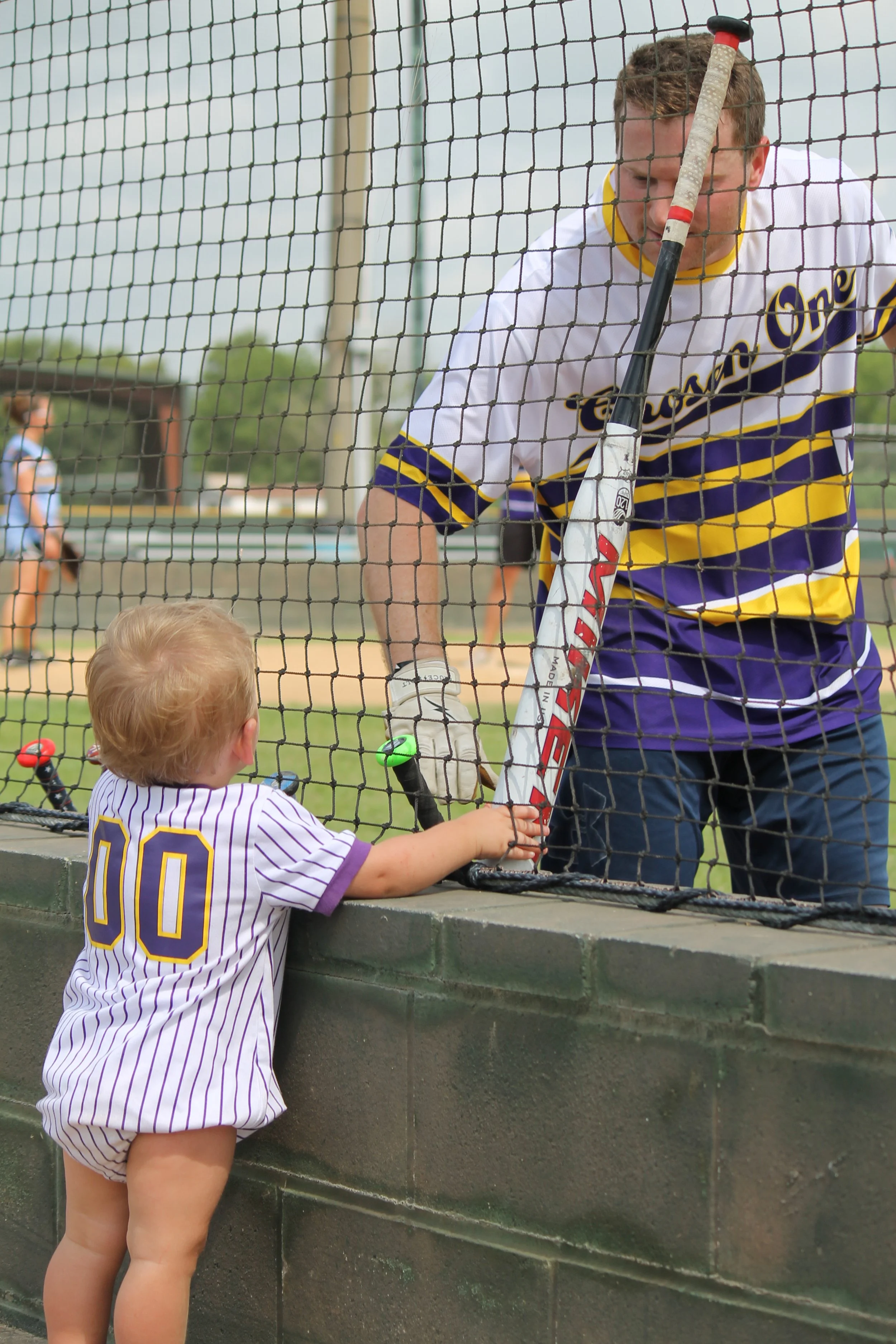 A young child in a striped baseball uniform reaching out to a man in a purple and yellow athletic jersey through a fence at a baseball field.