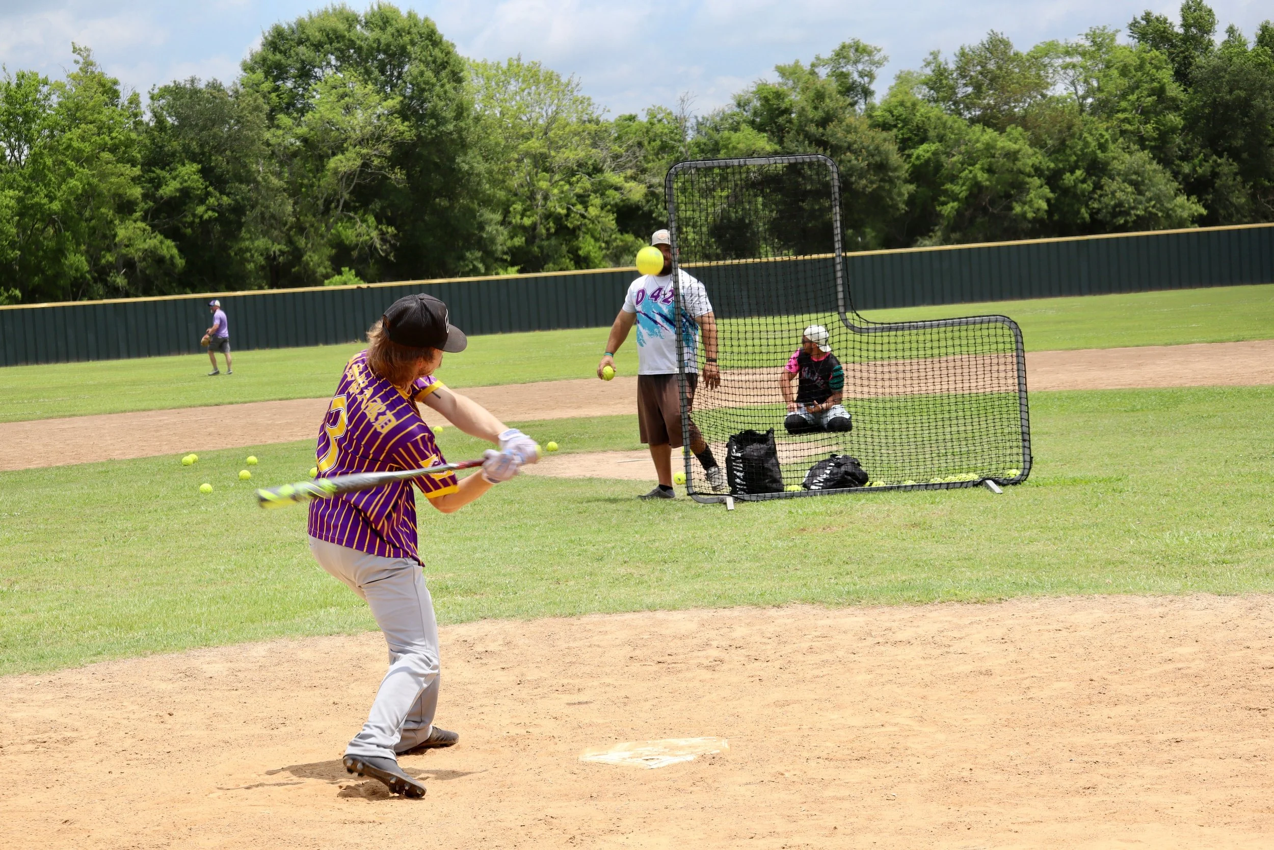 Person practicing baseball batting on a field with a youth baseball game ongoing in the background, including a person throwing a pitch and another near the batting cage.