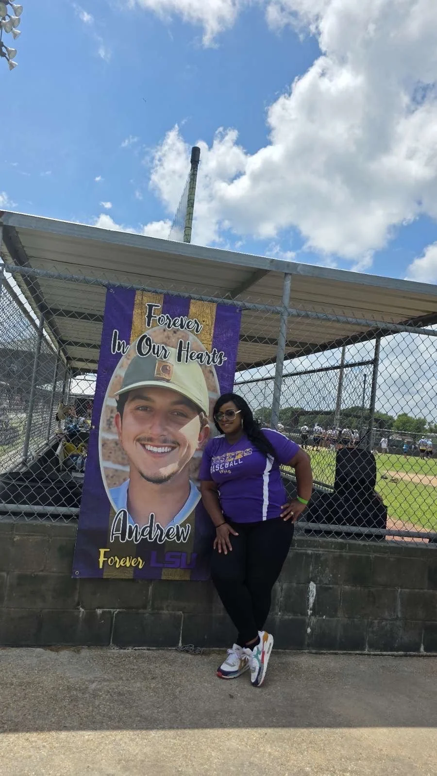 A woman standing in front of a purple LSU baseball banner with a smiling young man, at a baseball game under a partly cloudy sky.
