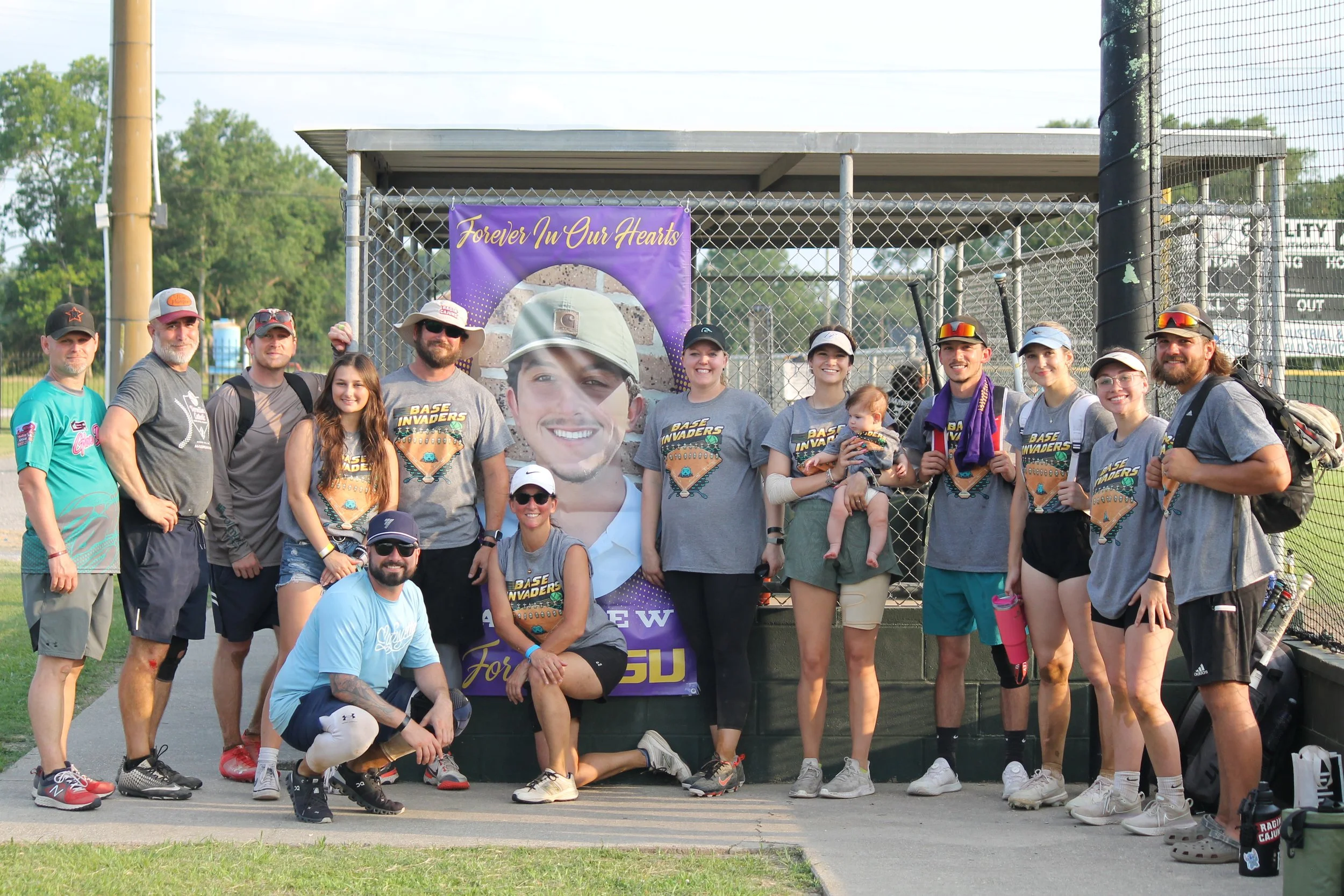A group of people posing in front of a chain-link fence with a purple banner that reads "Forever In Our Hearts." The group appears to be at a baseball field, with some wearing matching "Base Invaders" t-shirts and some holding a small child. The grou