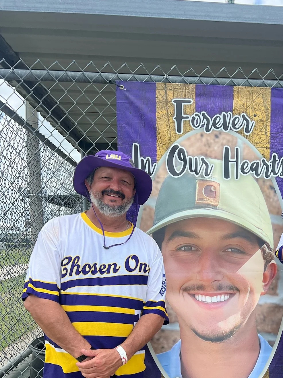 A smiling man wearing a purple LSU hat and a white and yellow LSU jersey standing in front of a banner that reads 'Forever In Our Hearts' with a large photo of a young man in military attire.