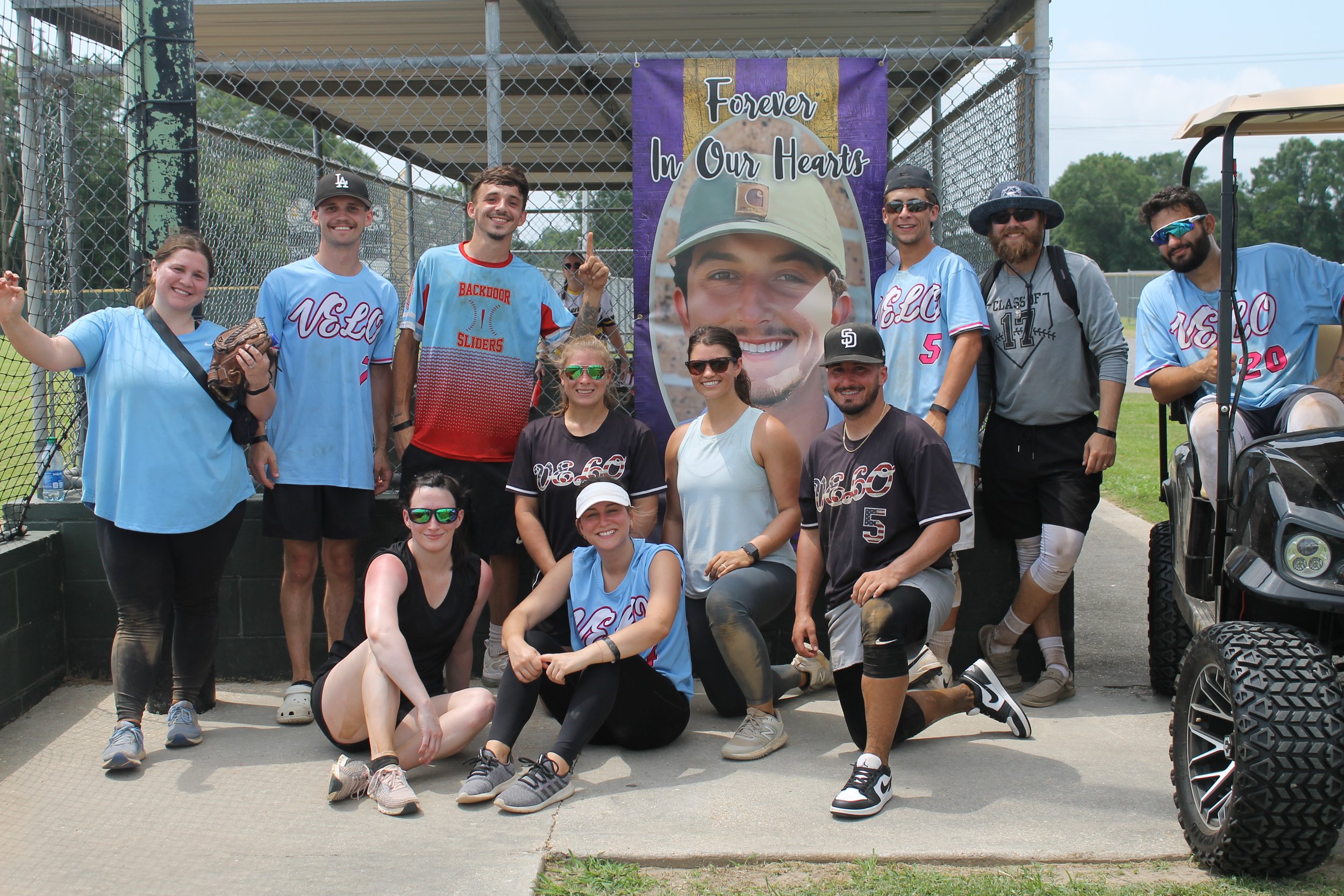 Group of people at a baseball field posing in front of a large memorial banner with a smiling young man in military uniform. Some individuals are kneeling and others standing, wearing casual sportswear, with a golf cart on the right side.