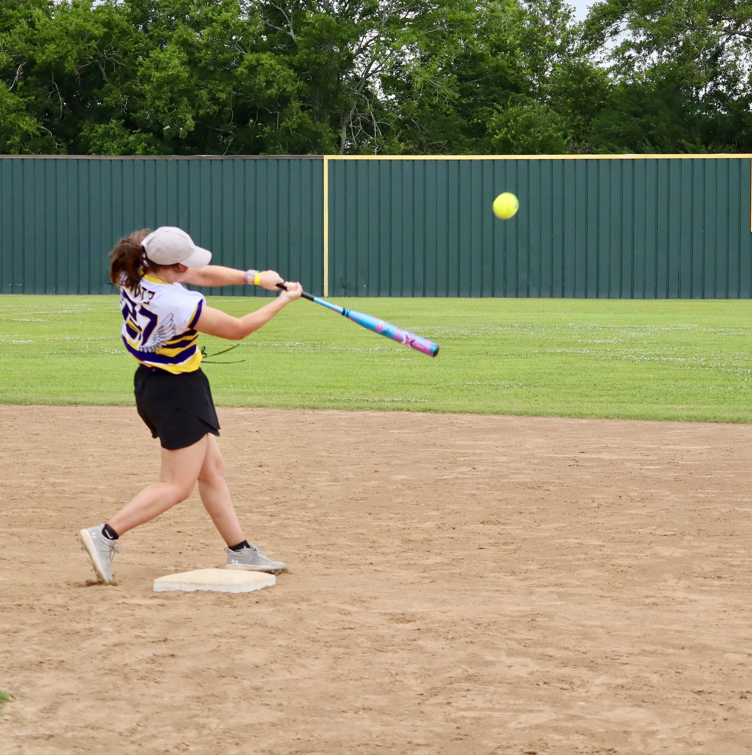 A girl playing baseball in a field, hitting a yellow-green ball with a colorful bat, wearing a baseball uniform, cap, and sneakers.