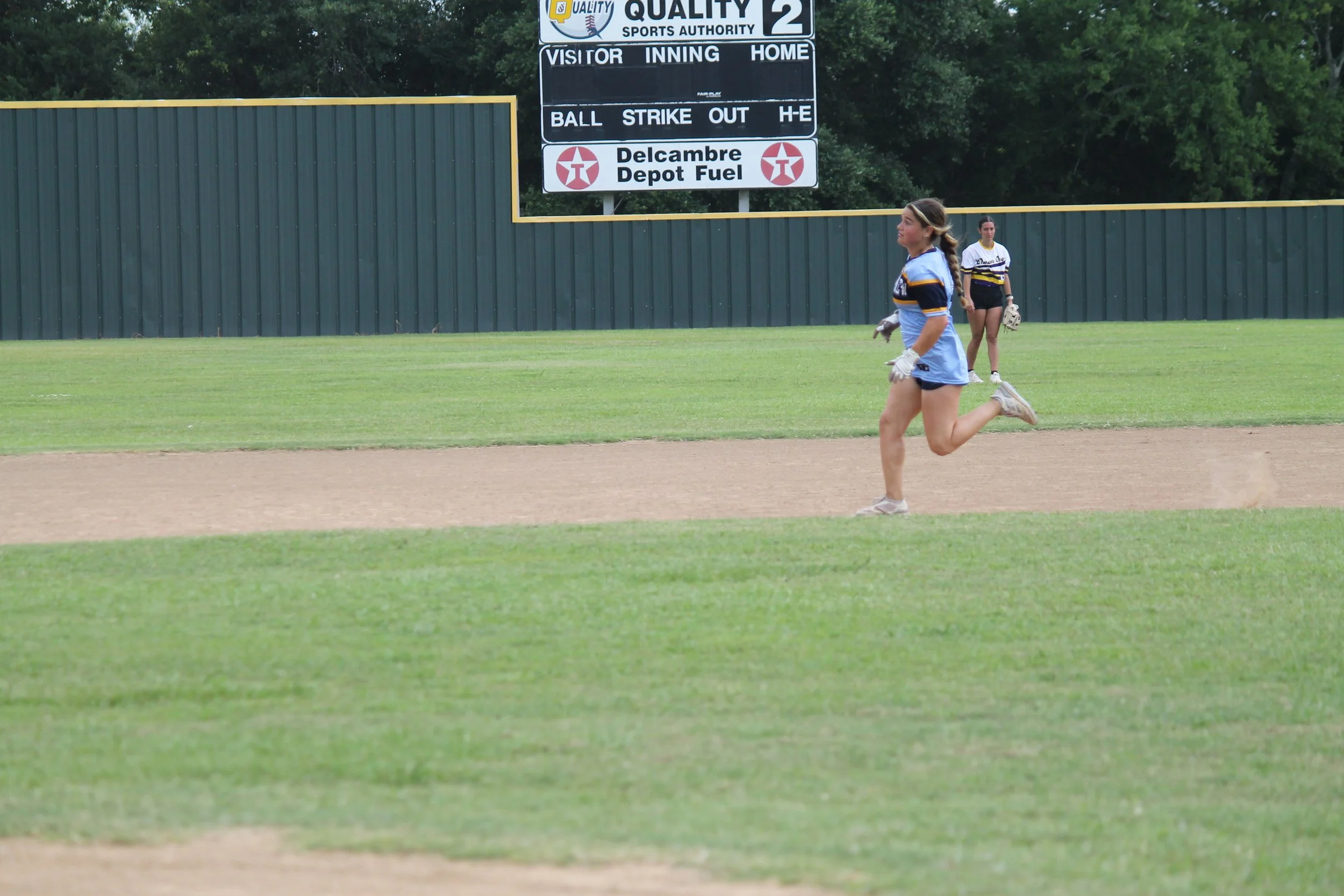 A girl in a blue and black sports jersey and shorts running on a baseball field during daytime, with another girl in similar attire standing in the background, and a scoreboard and green fence in the distance.