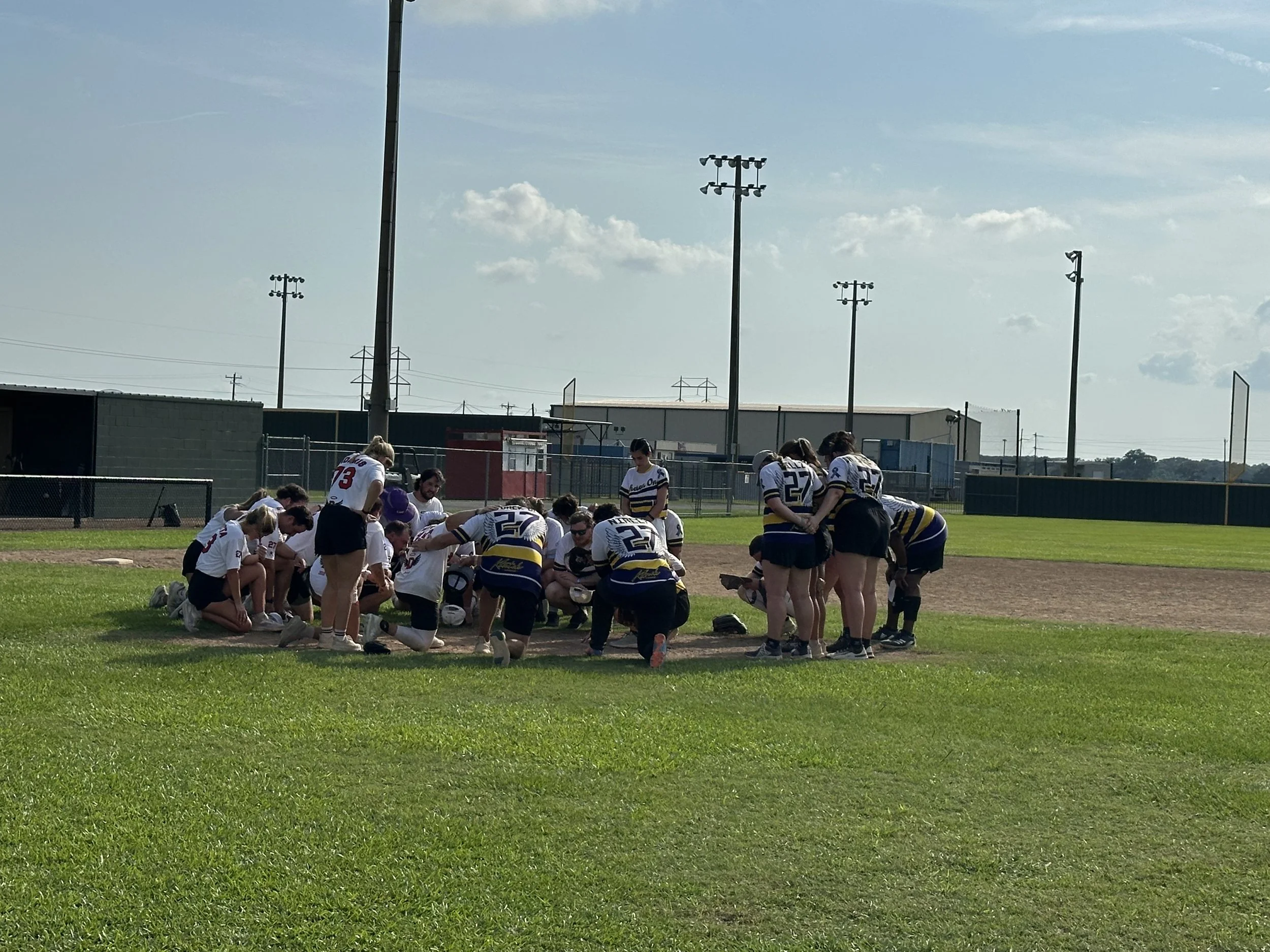 A group of female softball players wearing uniforms, gathered around their coach on a grassy field during daytime, with a baseball diamond, fence, and light poles in the background.