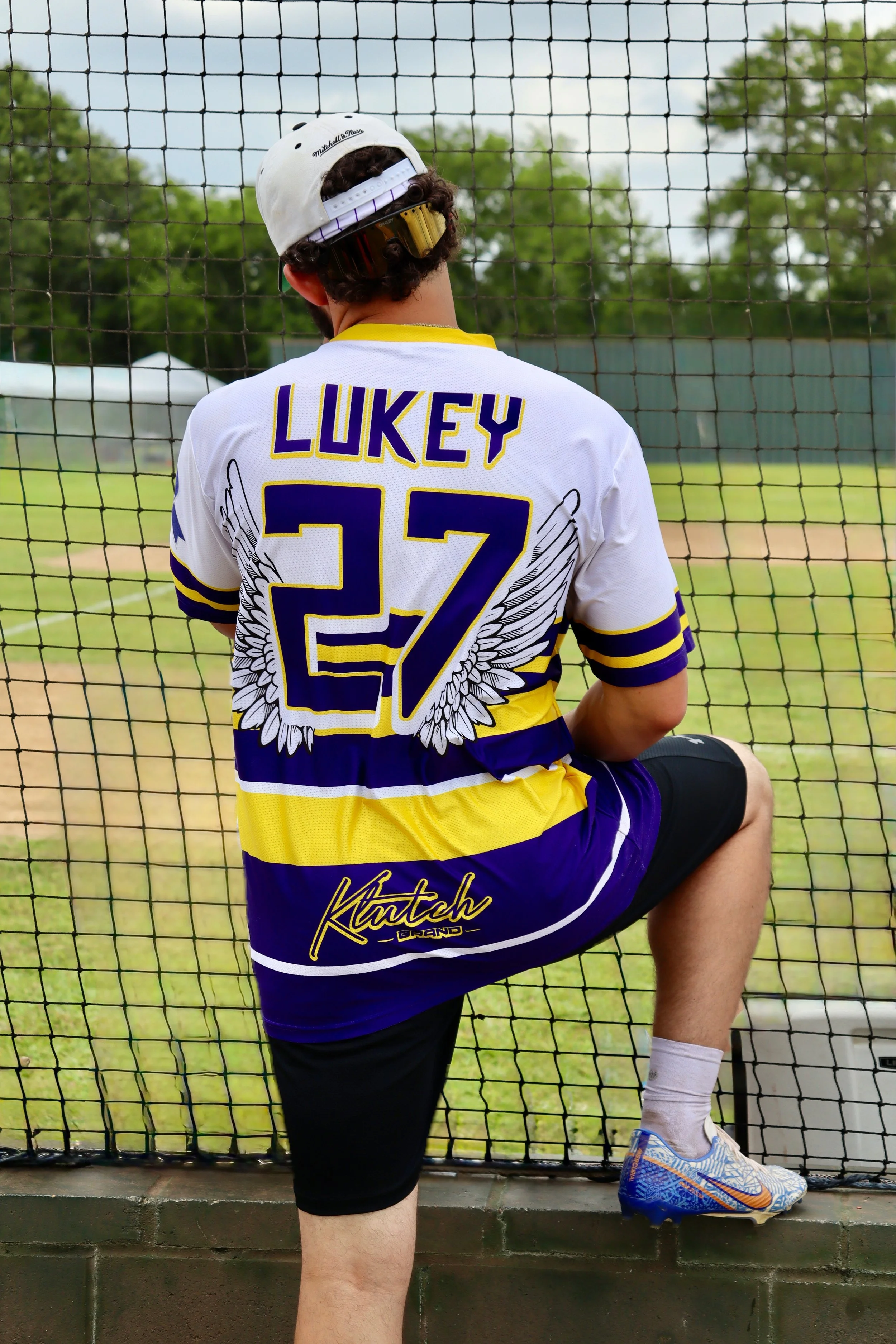 A male baseball player in uniform, sitting on the grass with one foot raised on a brick wall, looking through a net at a baseball field. The jersey has the name 'Lukey' and the number 27, with a winged design and the word 'Kutch' on the back.