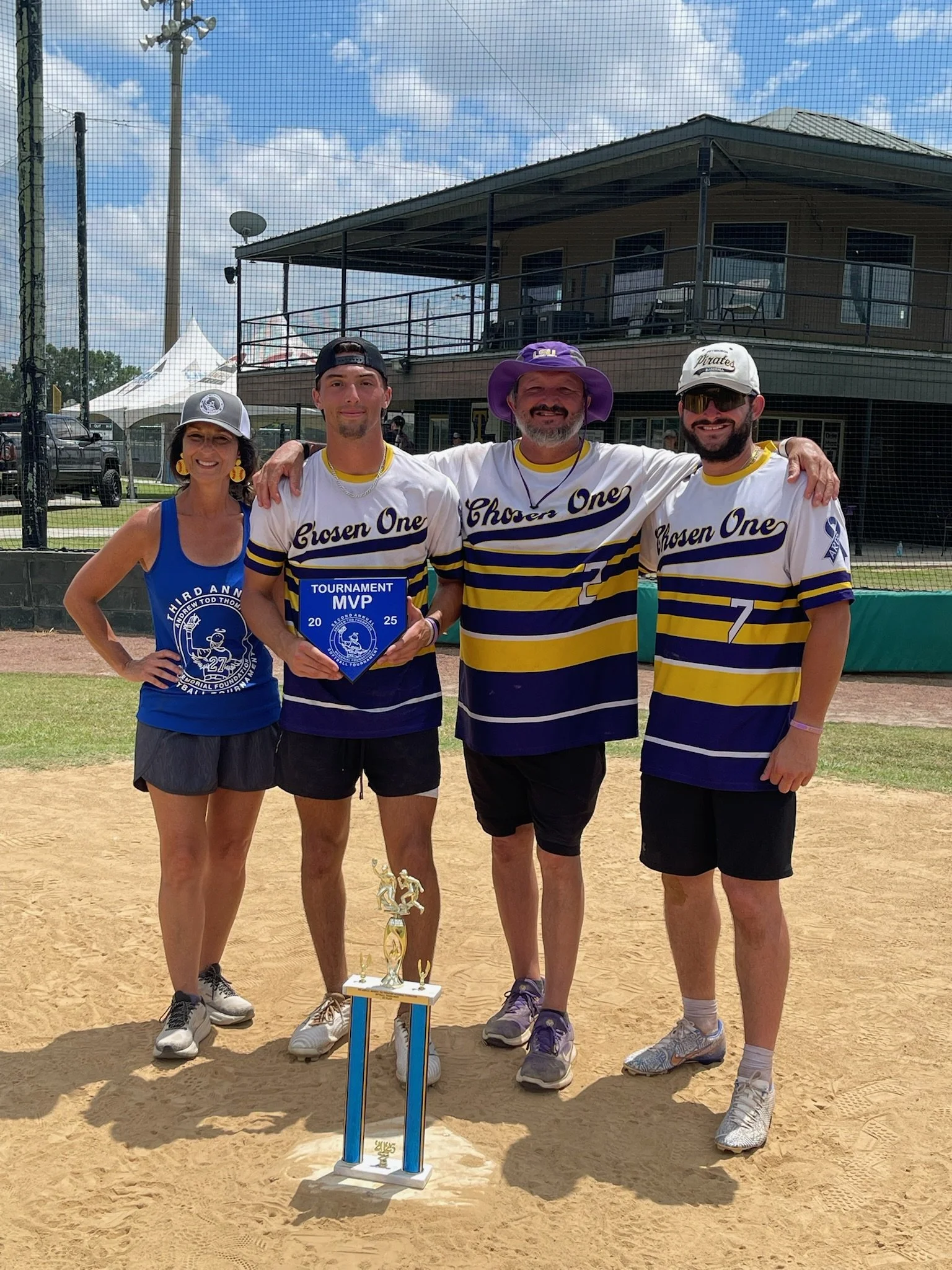 Four people standing on a baseball field celebrating a victory; the person second from the left is holding a blue tournament MVP plaque, and a tall trophy is on the ground in front of them. They are all wearing sports uniforms and have their arms aro