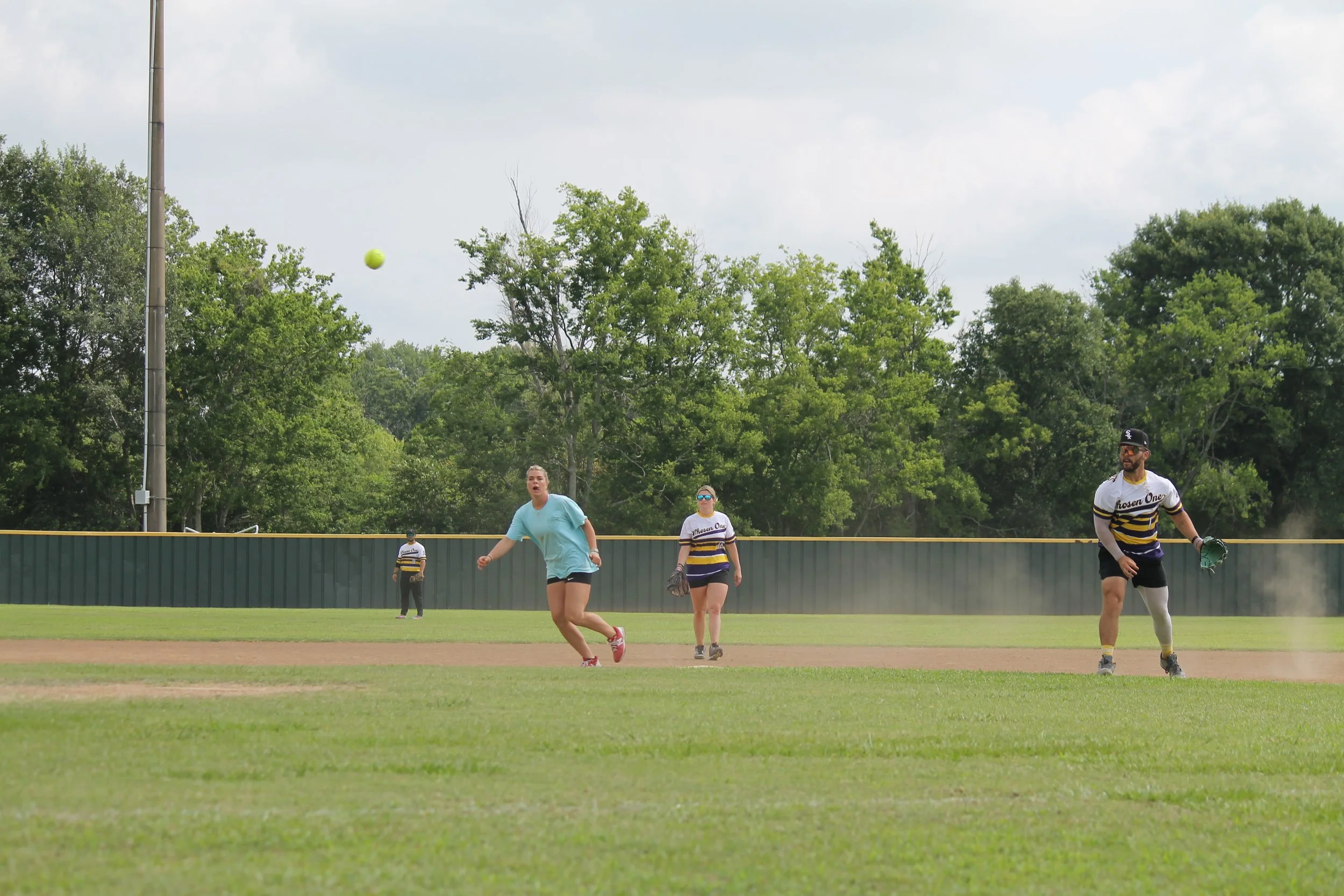 People playing baseball on a field with a grassy area and trees in the background.