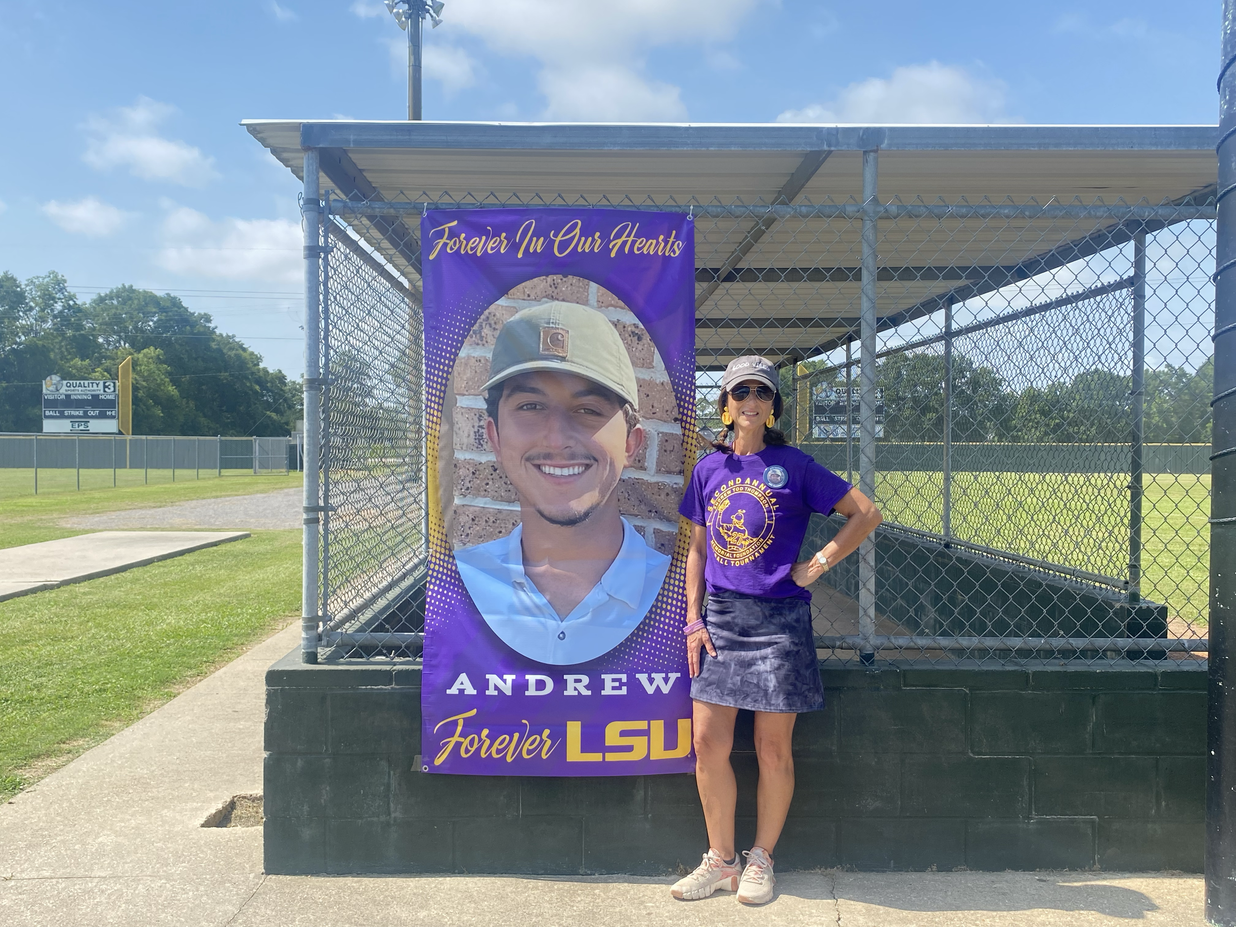 Woman standing next to a large poster of a young man in a baseball cap, with a sign reading 'Forever In Our Hearts'.