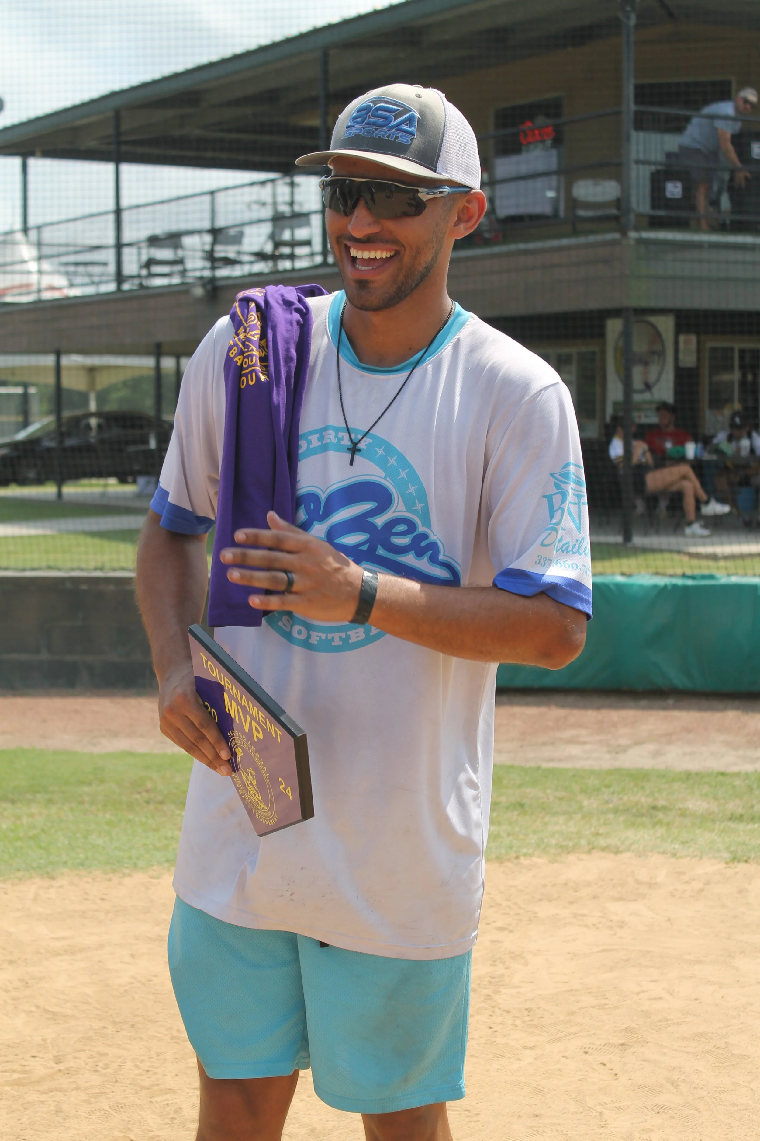 A man wearing sunglasses, a baseball cap, and a softball team shirt is smiling on a softball field, holding a trophy for a tournament MVP award.