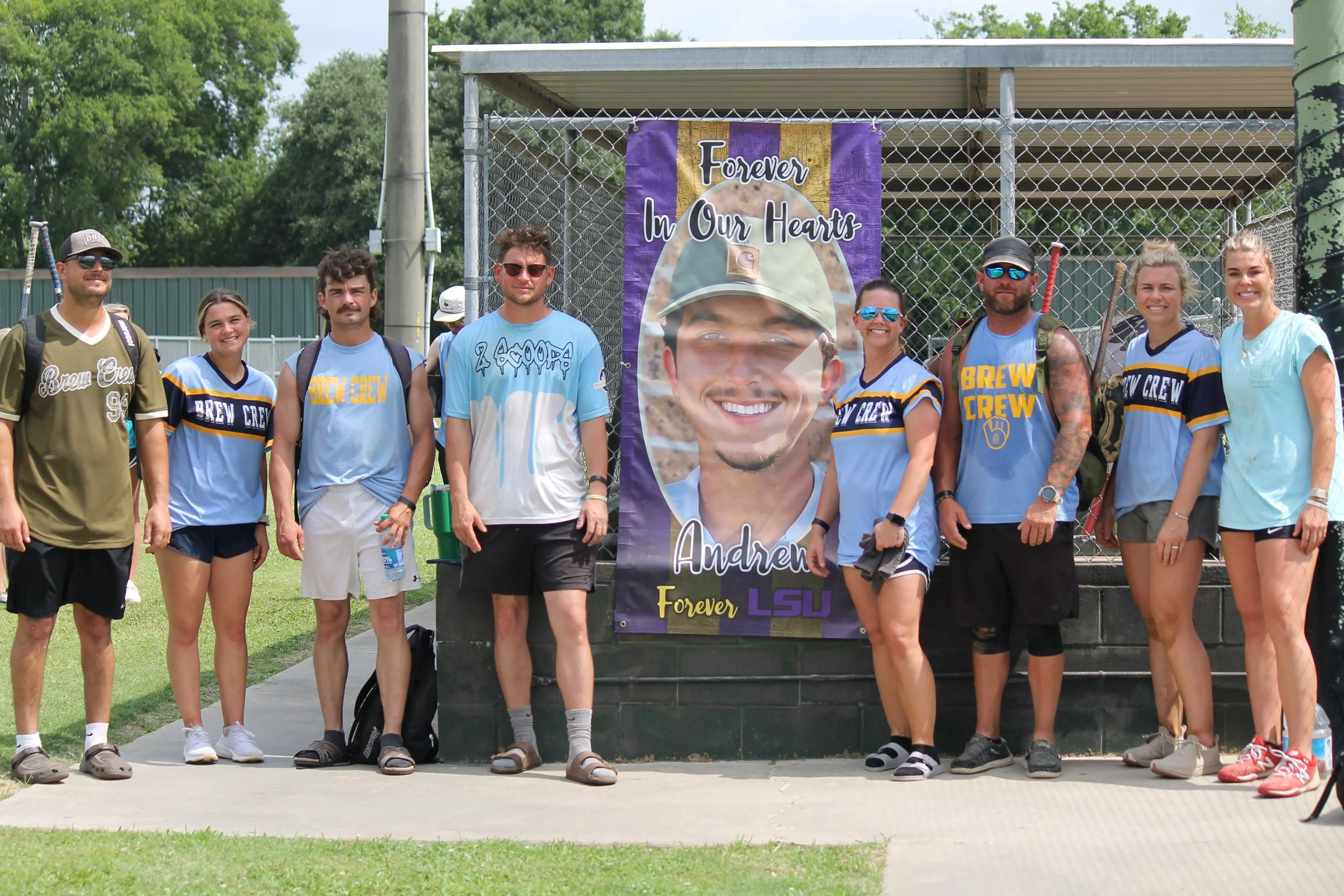 Group of eight people standing outdoors beside a large memorial poster that reads "Forever In Our Hearts Andrew Forever LSU". The group includes men and women wearing casual sportswear, some with backpacks, standing on a concrete pathway in front of 