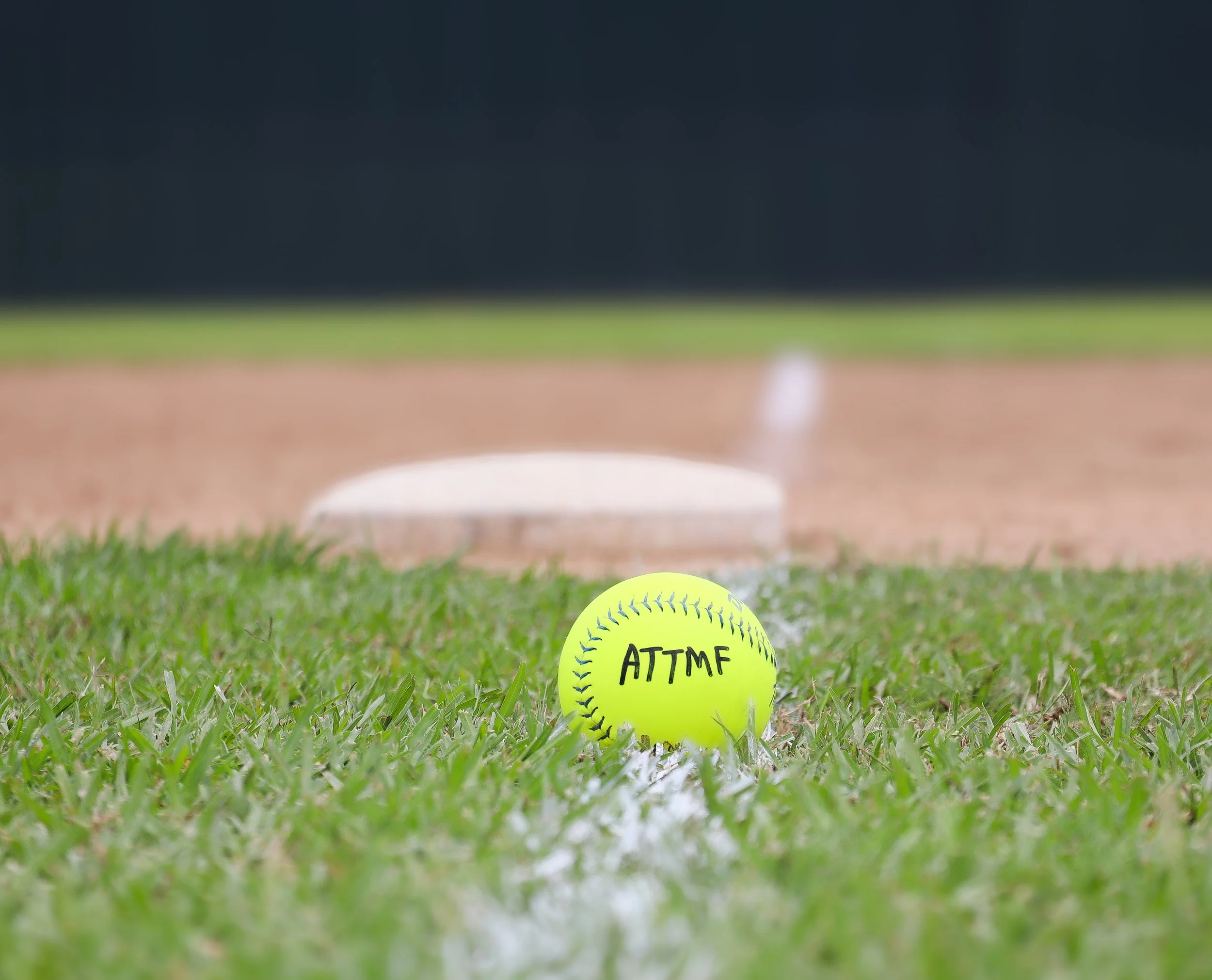 A close-up of a bright yellow softball on the grass with the handwritten word ATTMF on it, with a blurred softball in the background on the field.