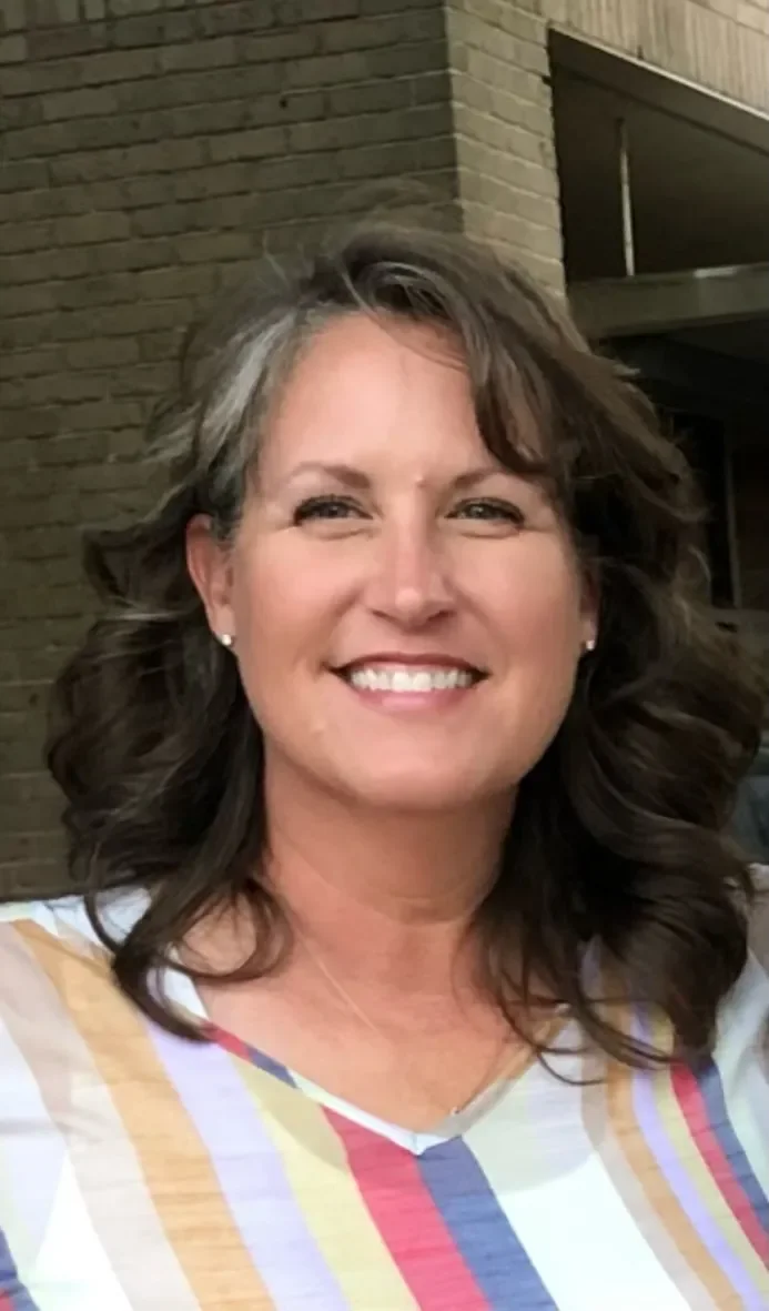 A woman with wavy, shoulder-length brown hair and a bright smile, wearing earrings and a colorful striped shirt, standing outside near a brick wall.