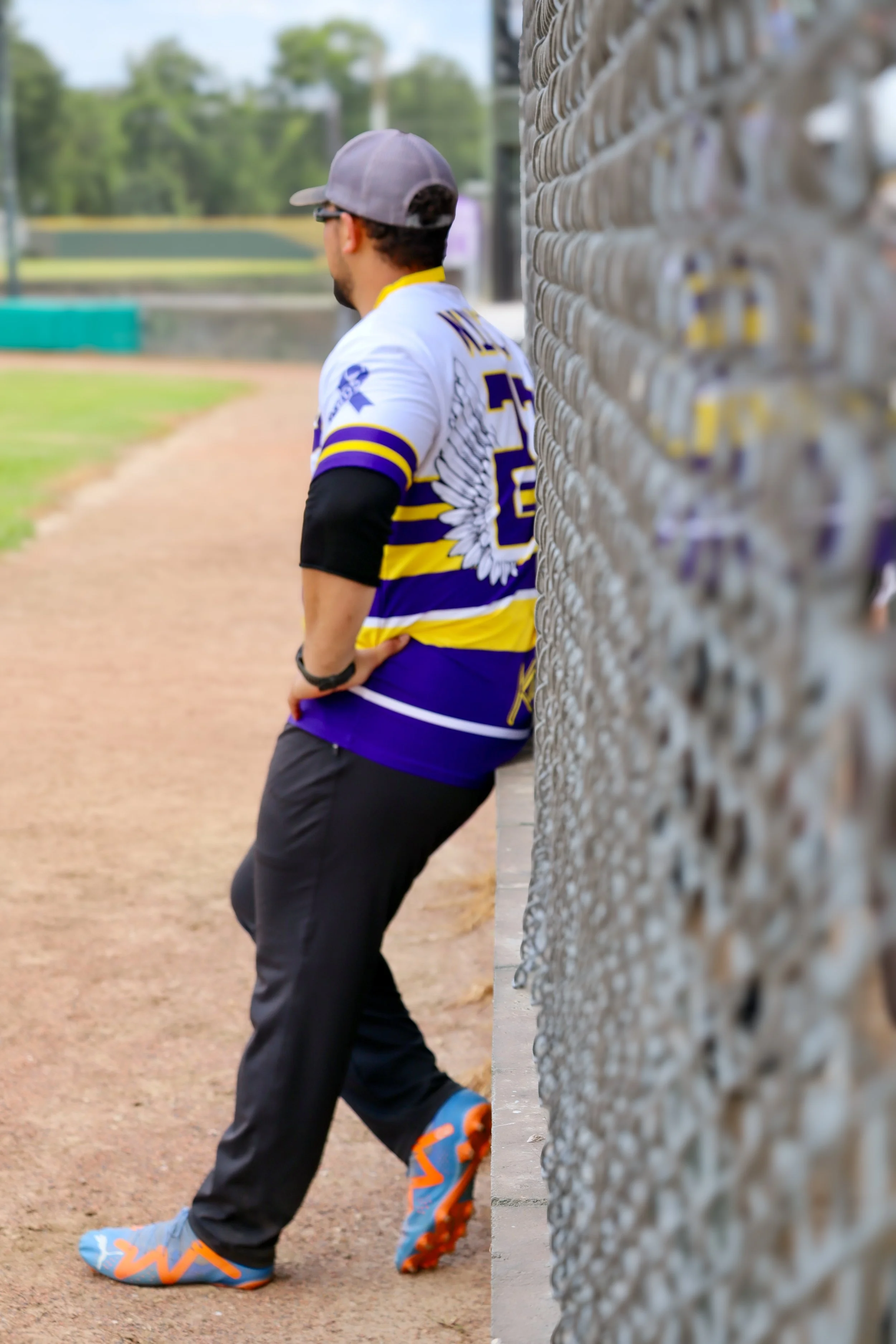 Man standing next to a chain-link fence, wearing a sports jersey, dark pants, and colorful shoes, on a baseball field.