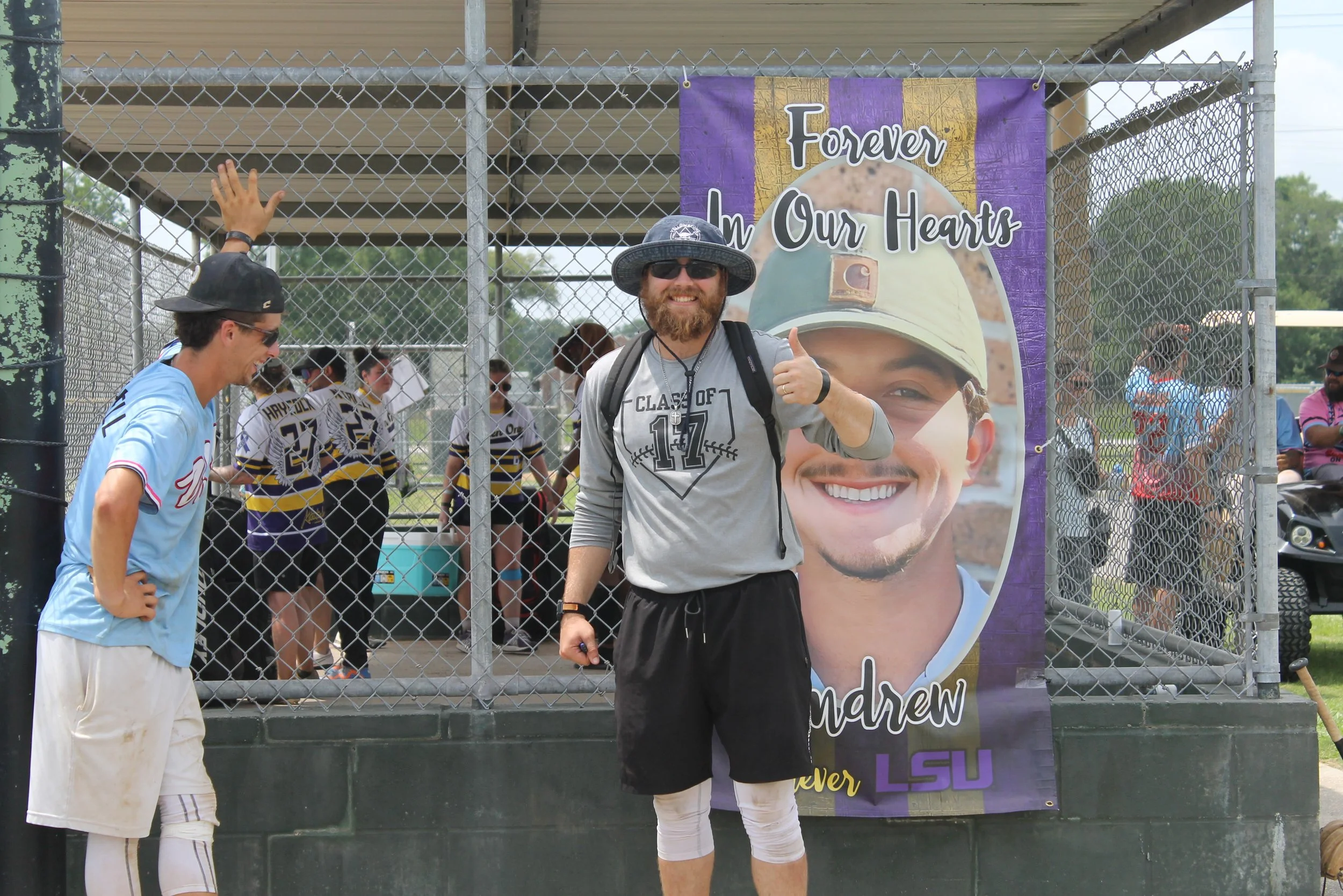 A smiling man with a beard, sunglasses, and a sun hat giving a thumbs-up in front of a large photo banner of a young man in military attire. The background shows athletes in sports jerseys and other people behind a chain-link fence, likely at a sport