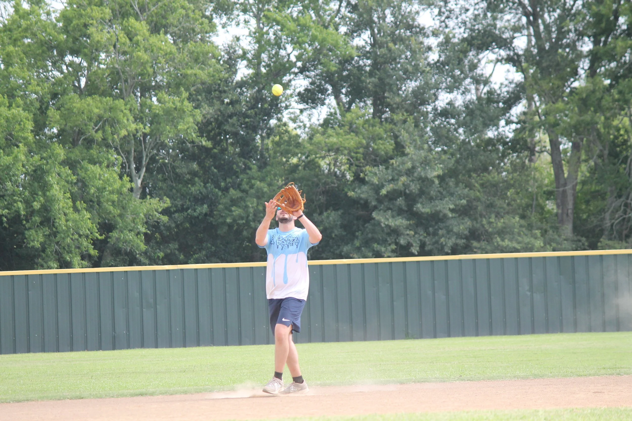 A young man playing baseball on a field, preparing to catch a ball with his glove, with a background of green trees and a gray fence.