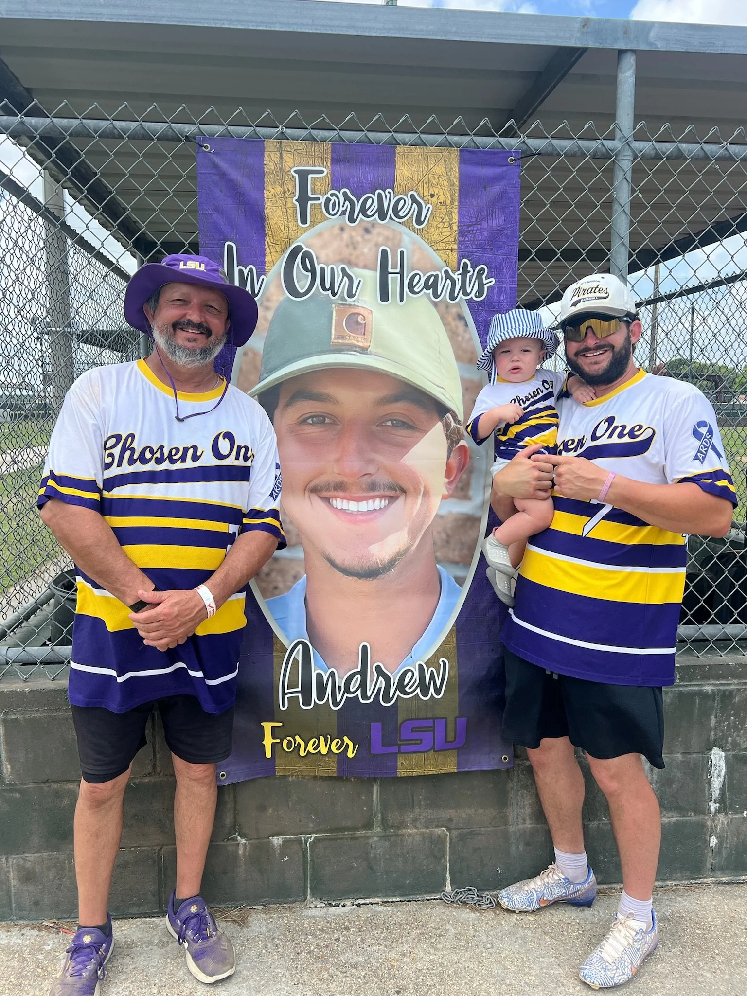 Man and boy wearing LSU baseball jerseys and hats standing in front of a memorial banner with a smiling young man's photo, with the words 'Forever In Our Hearts' and 'Andrew'. The man is wearing a purple LSU hat and the boy is holding a small child.