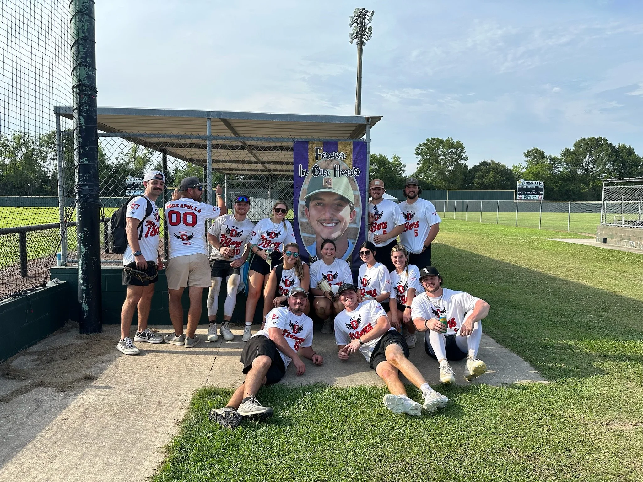 A group of ten baseball players in white jerseys with red and black accents pose for a photo in front of a large memorial sign with a picture of a smiling young man. They are on a baseball field with green grass and a chain-link fence in the backgrou