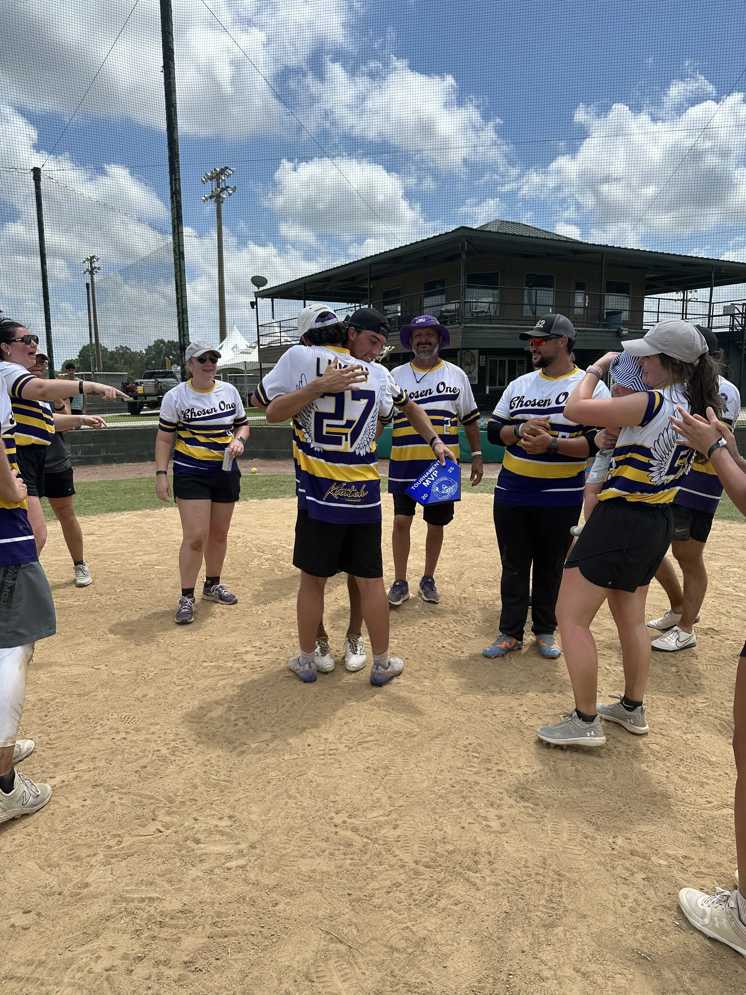 Group of people on a baseball field, some wearing matching jerseys, celebrating and smiling, with a man in the center wearing jersey number 27. Some people are pointing or touching the man, who is holding a blue folder or sign. The sky is partly clou