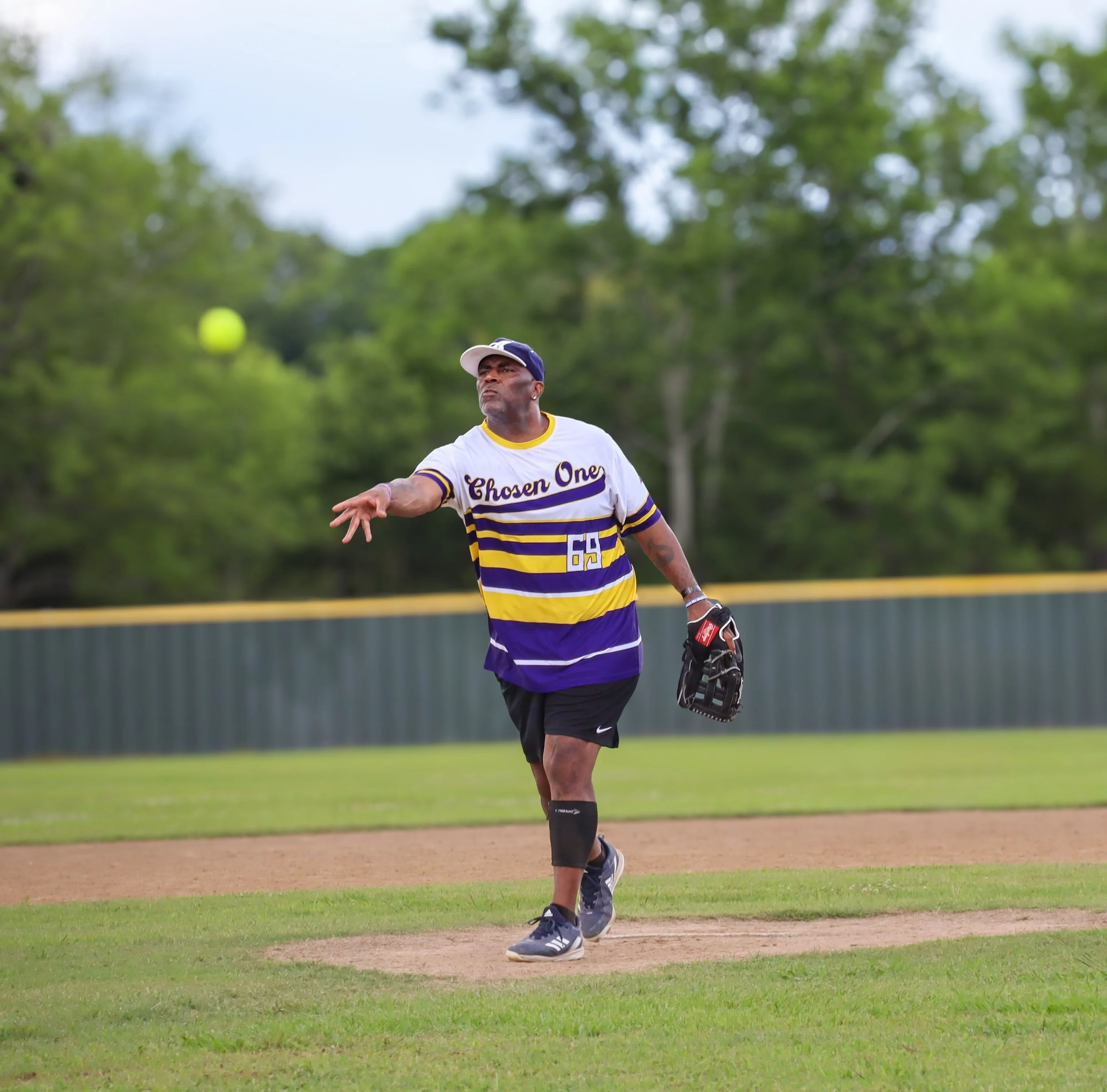 A man on a baseball field in the middle of a pitch, with a focused expression, wearing a white and purple striped jersey that says 'Chosen One' and the number 55, and holding a baseball glove in his left hand, with a green fence and trees in the back