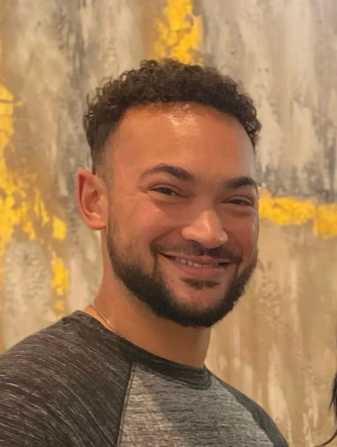 A smiling man with curly dark hair and a beard, wearing a gray and black shirt, standing in front of a curtain with yellow and gray patterns.