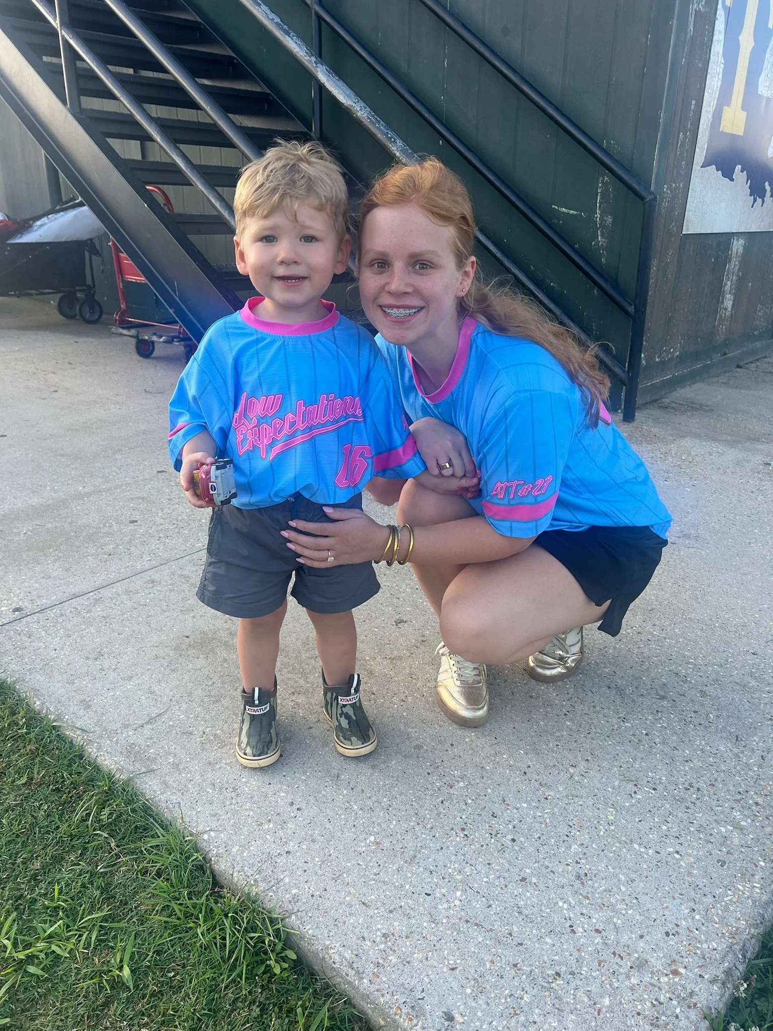 A young boy and a woman, possibly his mother, wearing matching blue sports jerseys with pink accents and text, smiling outdoors near a building with a staircase. The boy has blonde hair and is holding a small toy or device, while the woman has long r