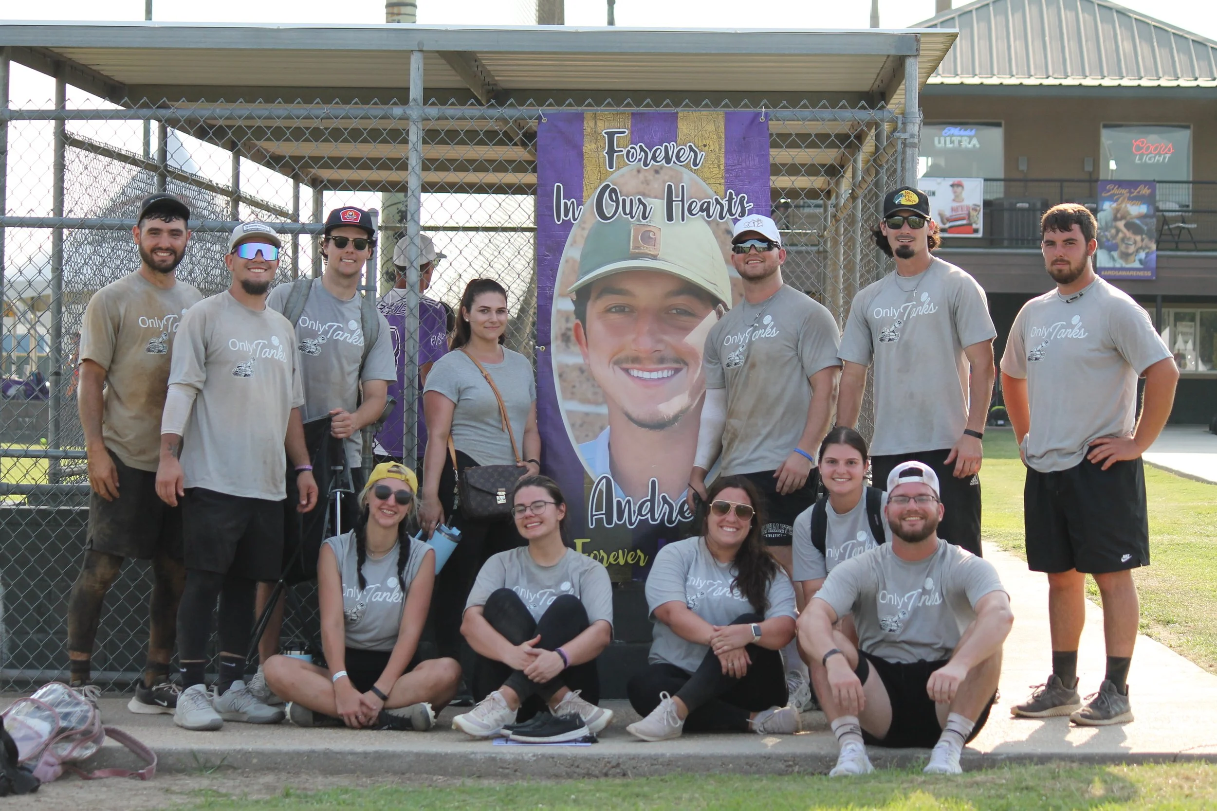 Group of people posing in front of a memorial banner with a smiling young man in a military helmet, with the text "Forever In Our Hearts" and "Andrew Forever." The group includes both men and women, some wearing gray shirts with "Only Tanks" printed 