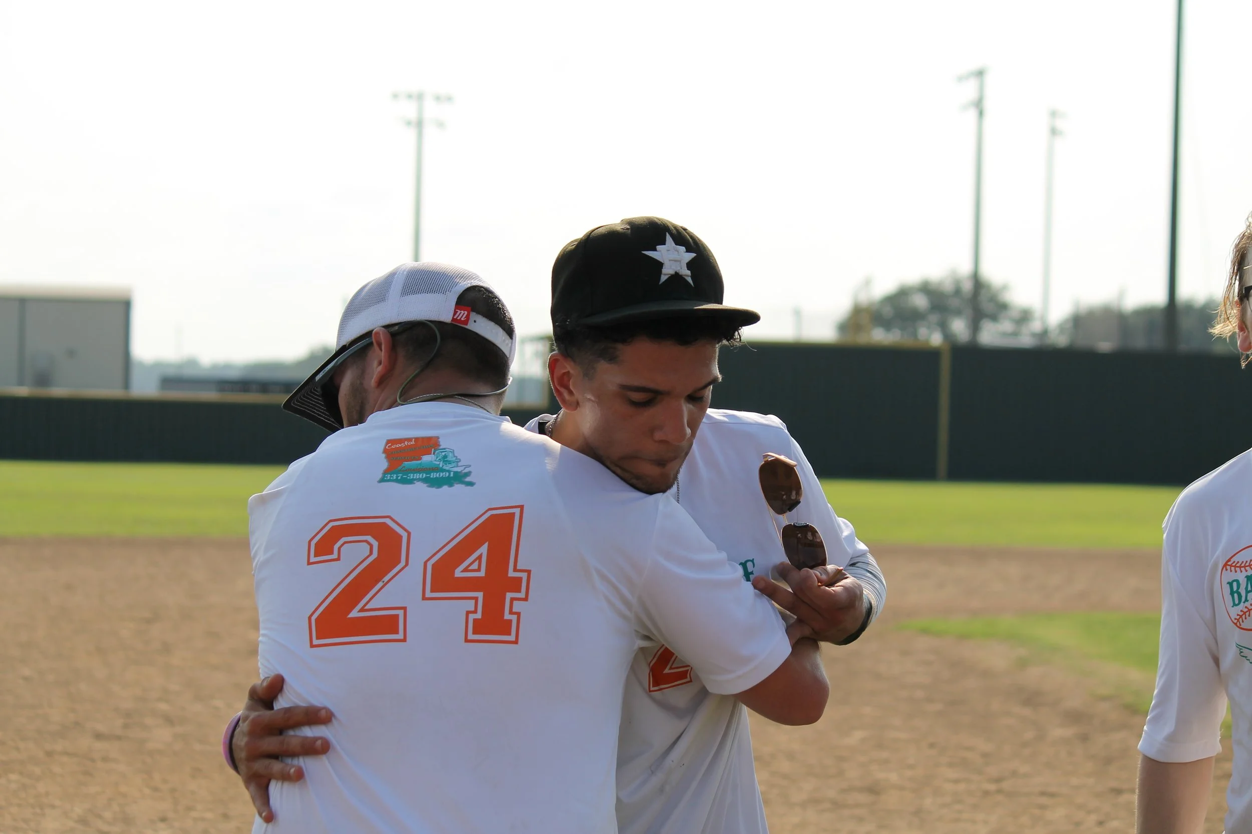 Two young baseball players hugging on the field, with one holding sunglasses, in front of a baseball field with a green fence and tall lights.