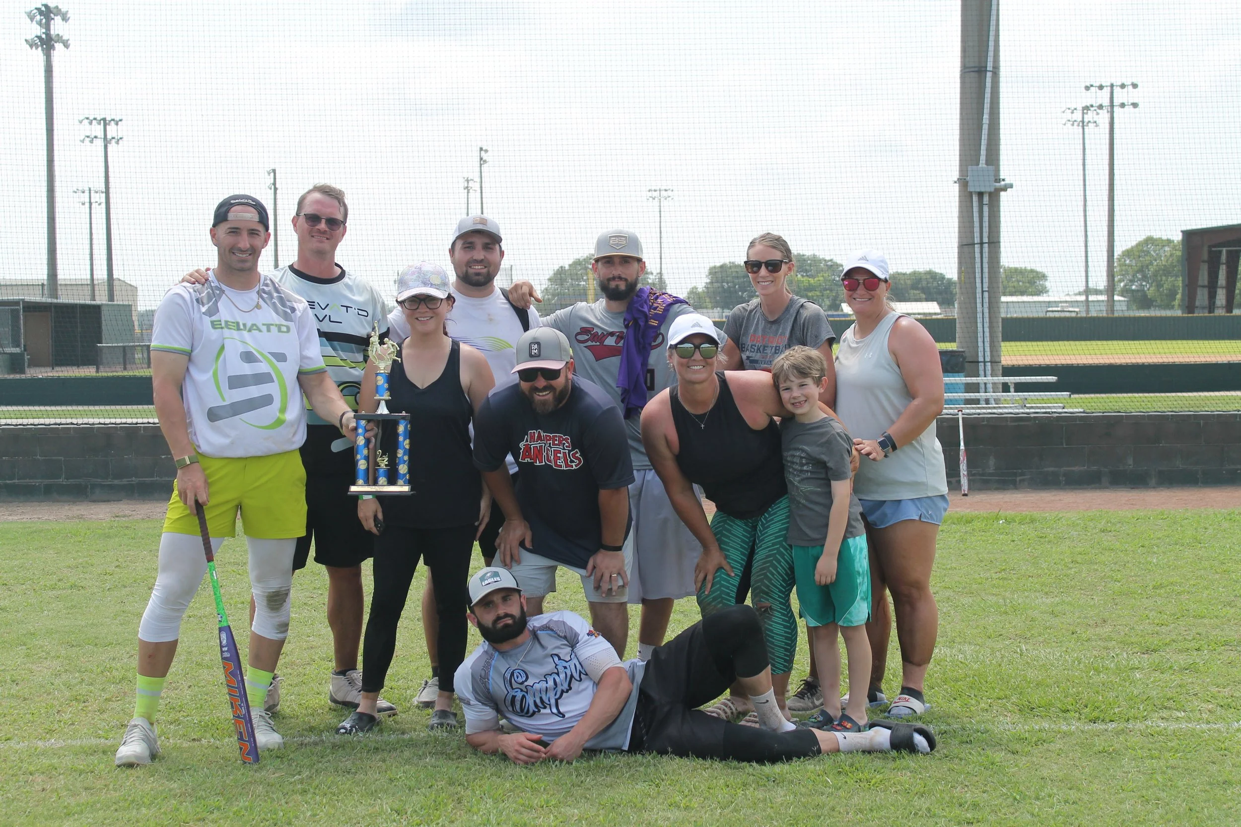 Group of people, including children and adults, posing outdoors on a grassy field with baseball and softball equipment. Some are holding a trophy, and they appear to be celebrating a sports victory or event.
