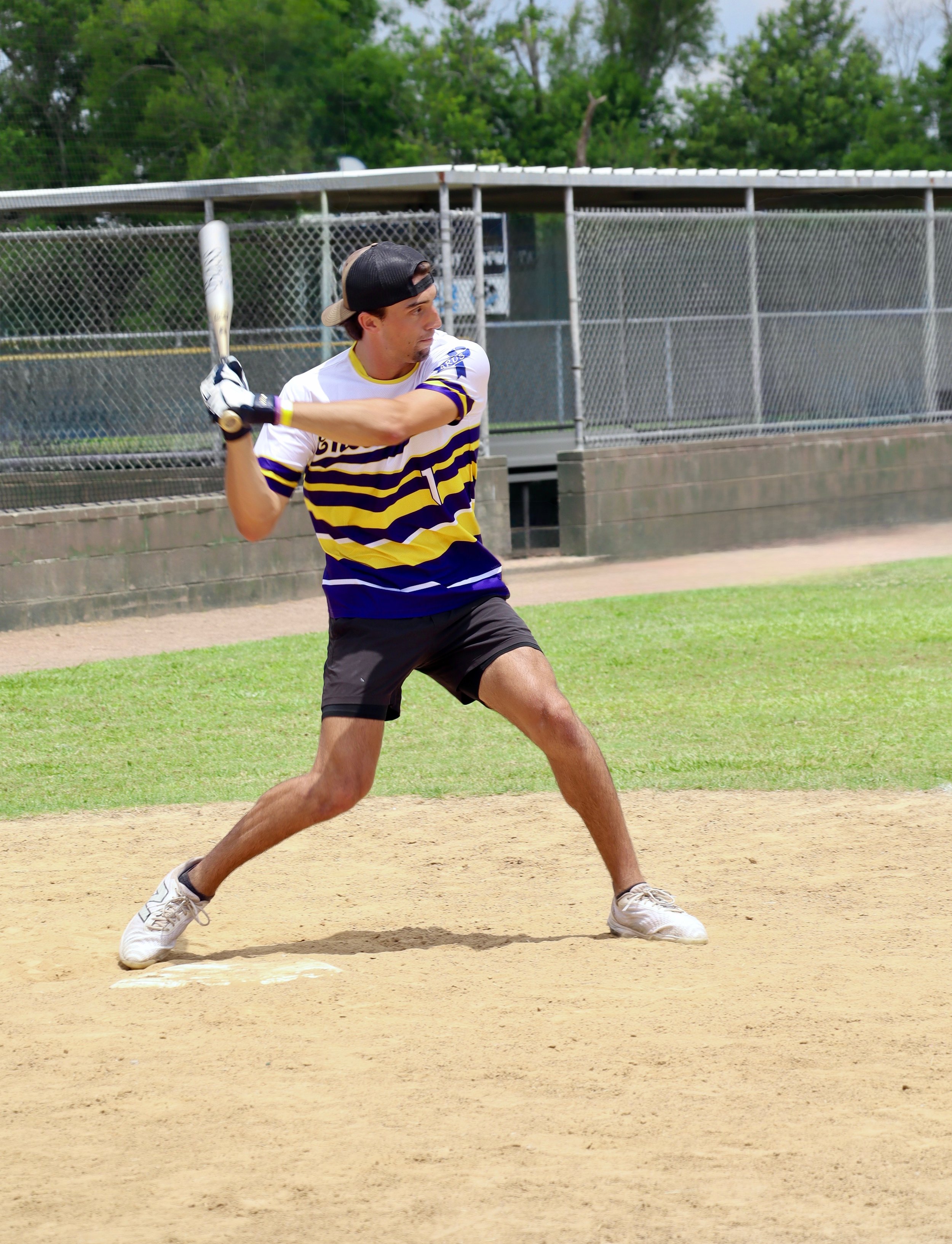 A young man playing baseball or softball at a ballpark, holding a bat in a ready stance on the dirt field with green grass and fencing in the background.
