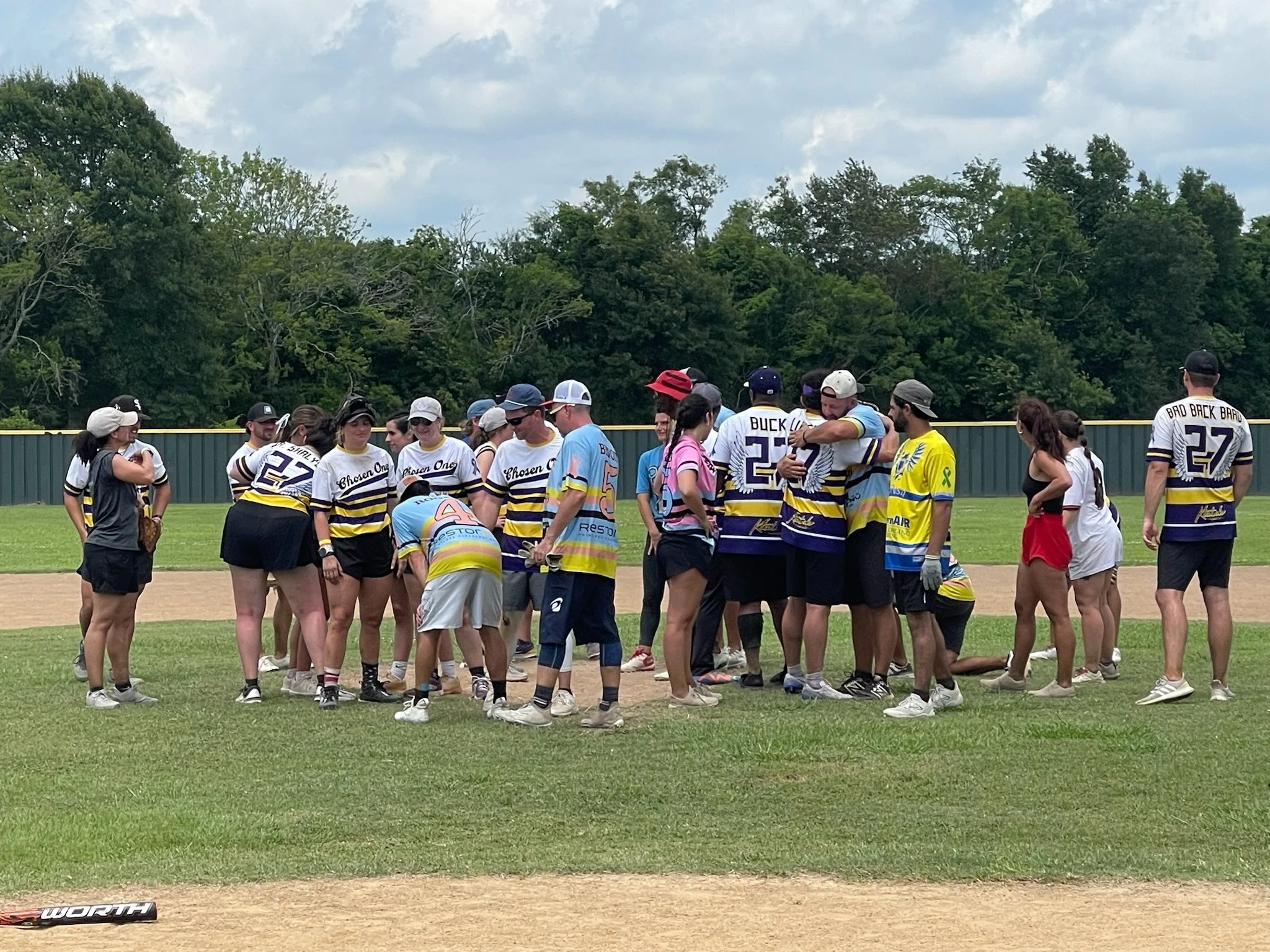 A group of softball players gathered on the field in a huddle, with some standing and others kneeling, wearing team jerseys and caps, during a game under a partly cloudy sky.