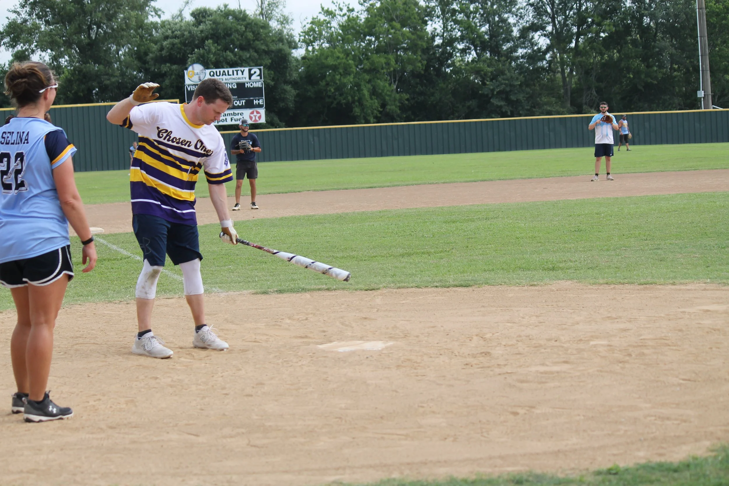 A softball player prepares to bat while standing on home plate, with a teammate and several players in the background at a baseball field.