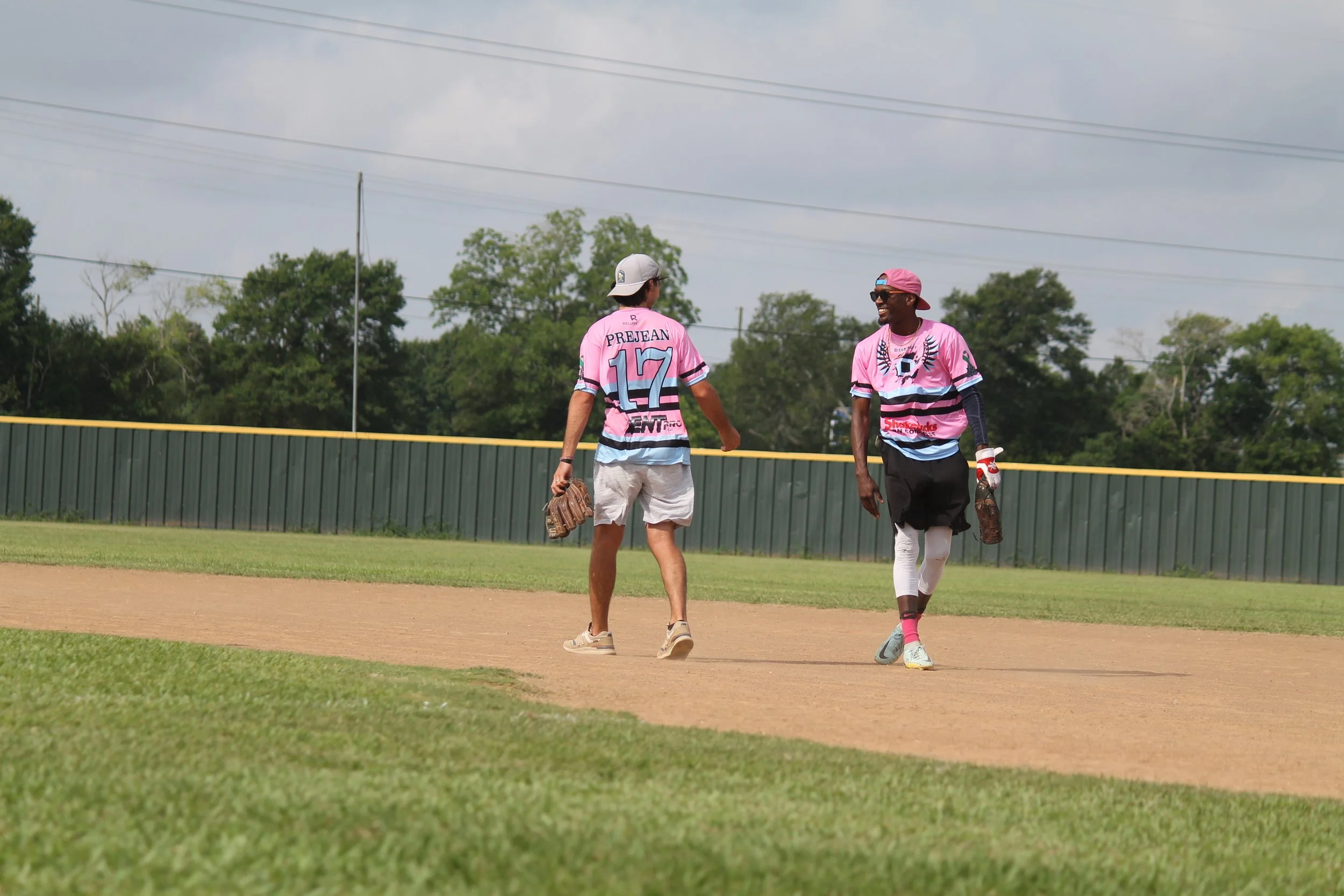 Two baseball players wearing pink jerseys walking on the field, with green grass, a dirt infield, and a green fence in the background. One player is wearing shorts and a cap, while the other is wearing long leggings and a cap, and they are smiling an