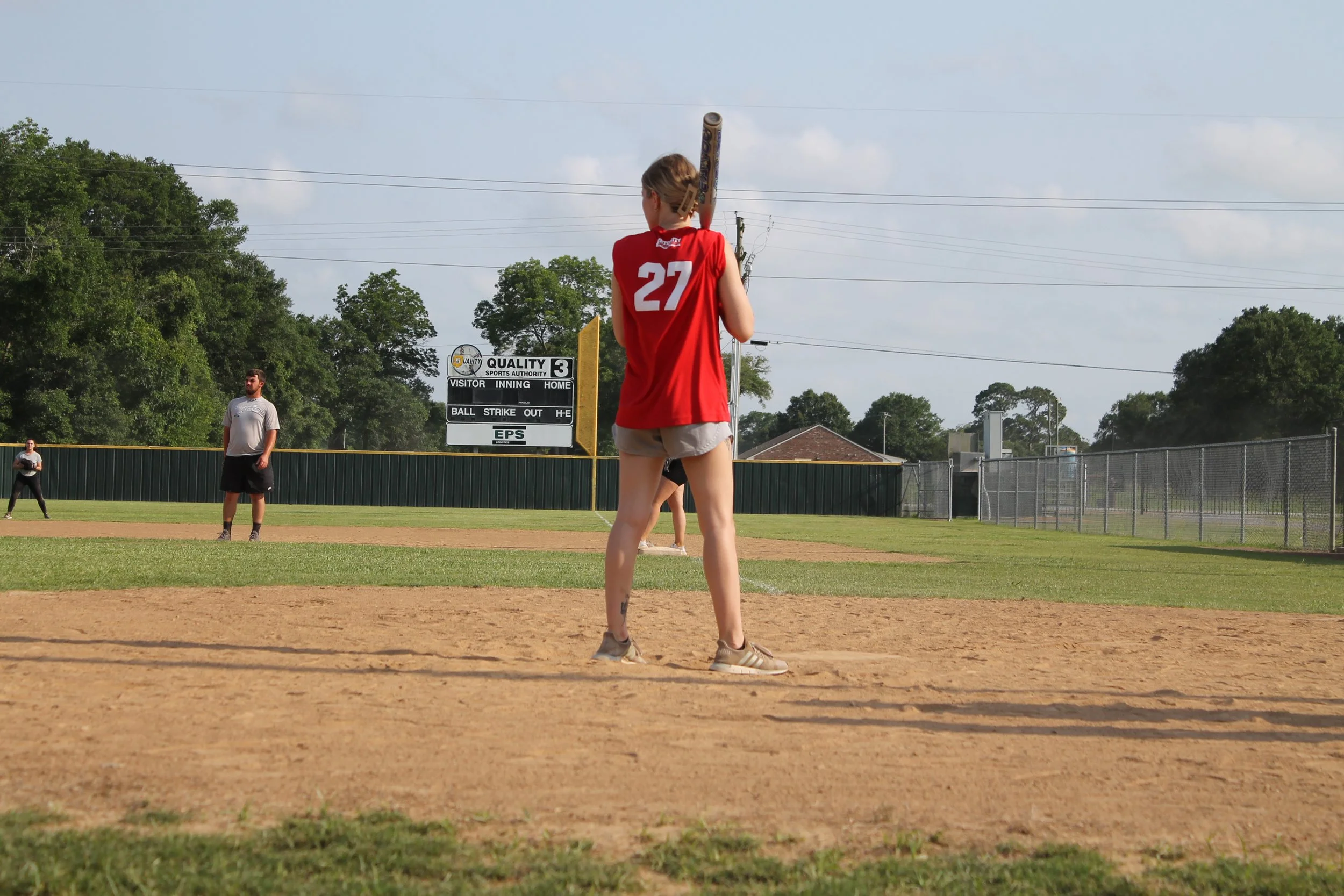 A young woman in a red sports jersey with the number 27 holding a baseball bat, standing on a baseball field. Other players are in the background, and the weather appears sunny and warm.