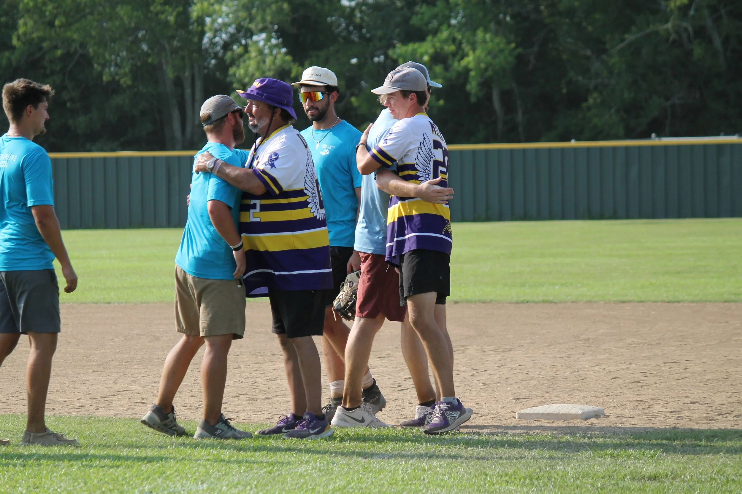 Group of men on a baseball field hugging and smiling, with others observing, during daylight