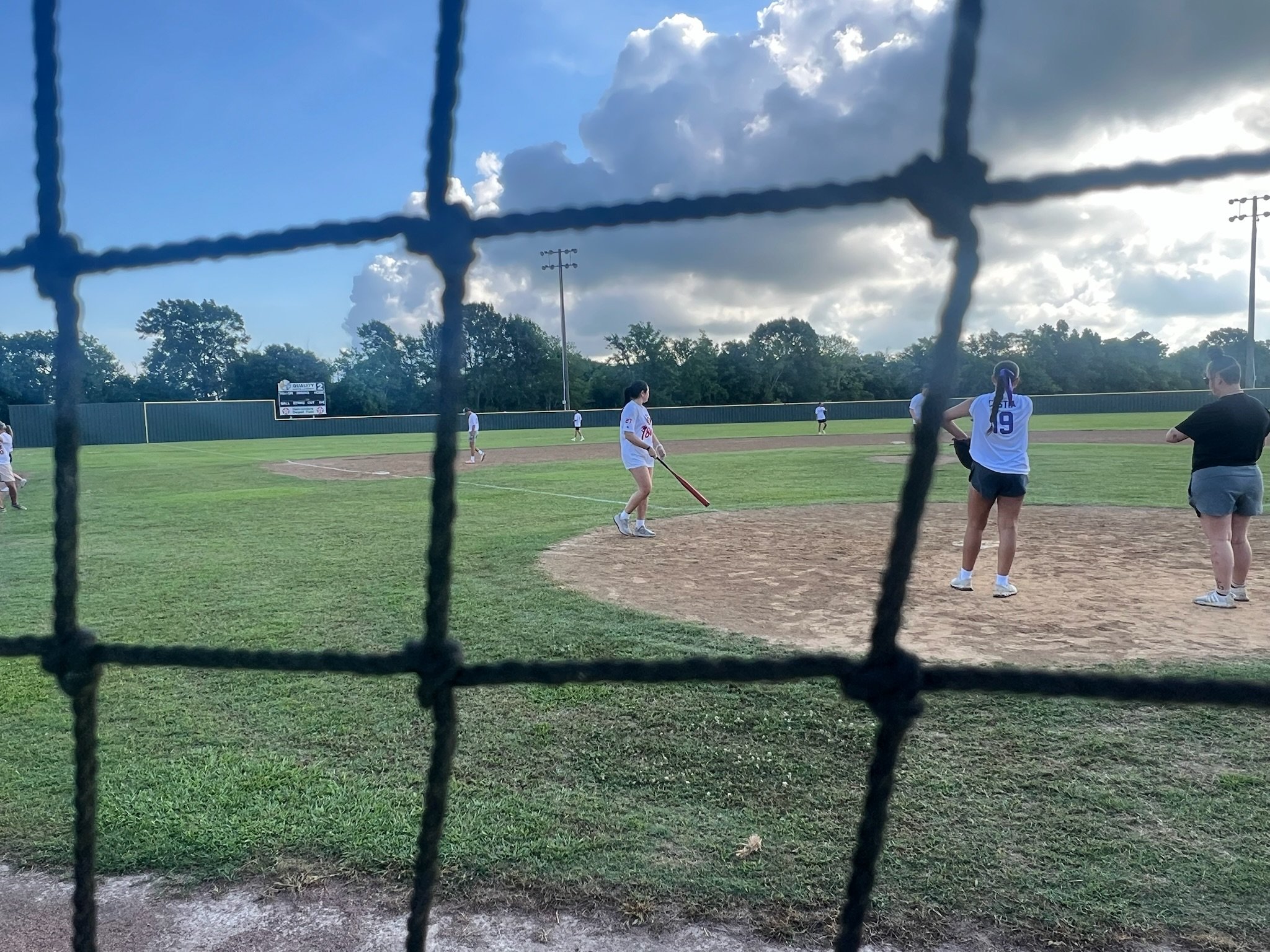 A girls' softball game as viewed through a chain-link fence on a partly cloudy day, with players on the field, some standing near the pitcher's mound and others in the outfield.