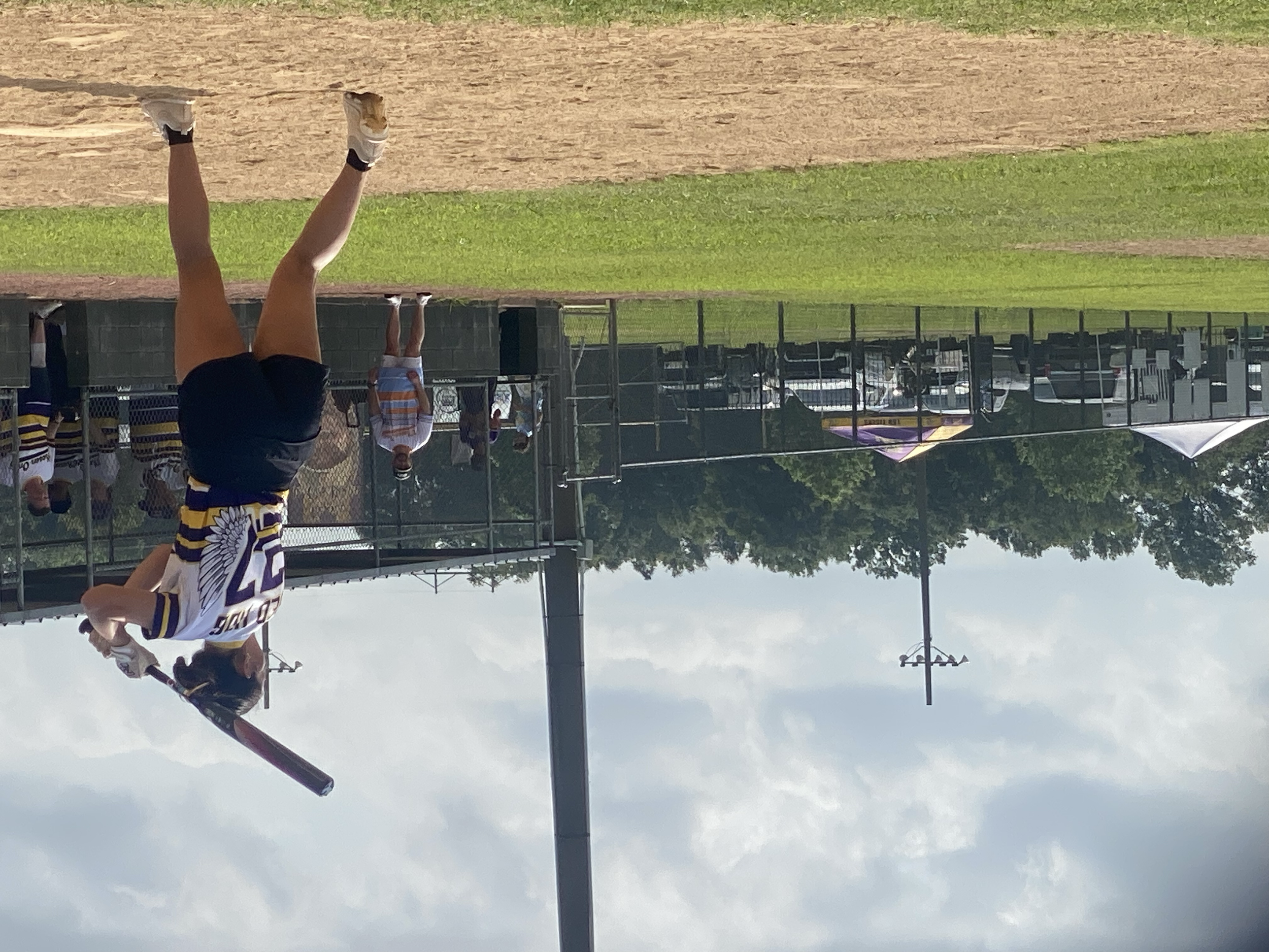 A girl in a sports uniform holding a bat, walking on a baseball field, with other players and spectators in the background.