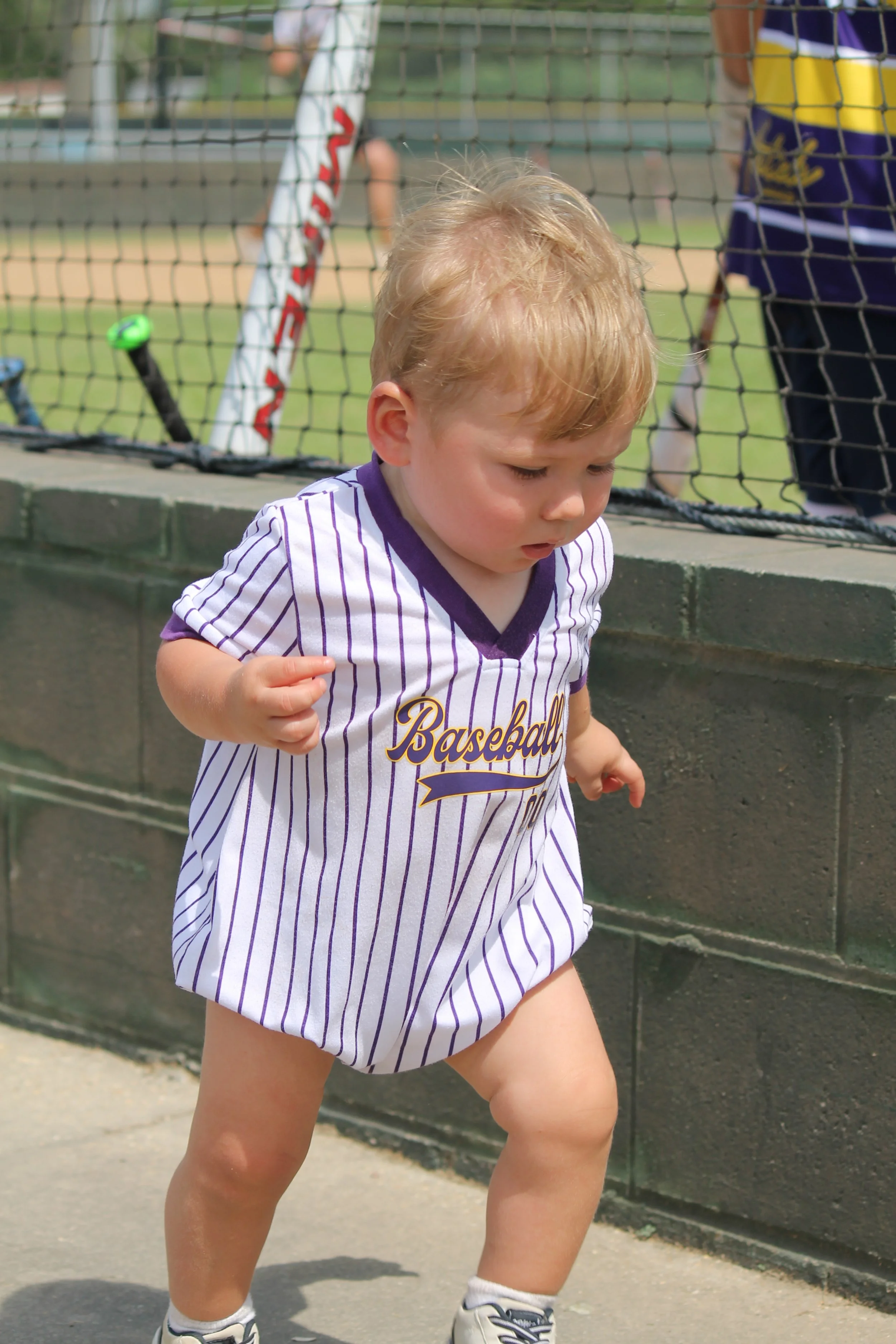 A young child wearing a white and purple striped baseball jersey running outdoors near a sports field.