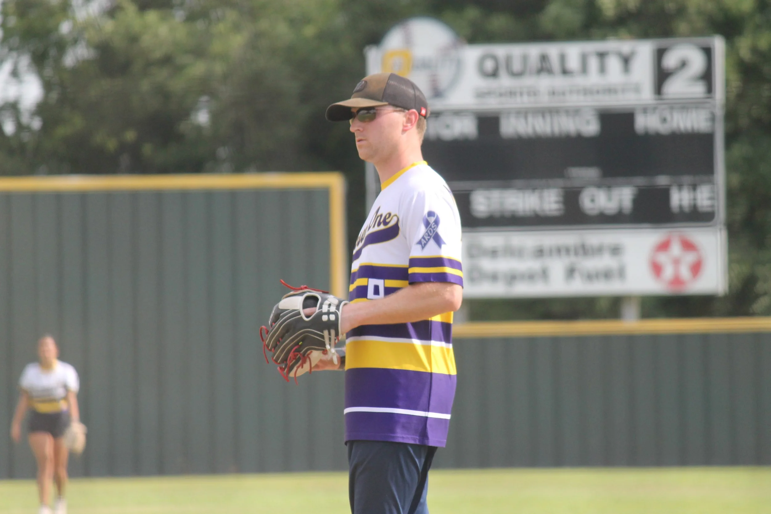 A baseball player standing on the field, wearing sunglasses, a cap, and a striped jersey, holding a baseball glove in his left hand, with a scoreboard and another player in the background.