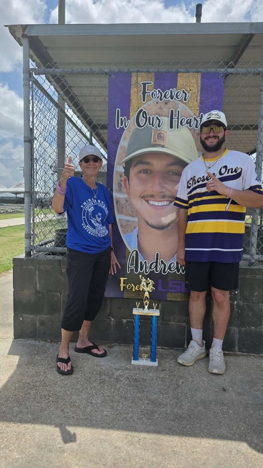 A woman and a man standing next to a memorial poster of a young man wearing a baseball cap. The woman is giving a thumbs-up, and the man is pointing at the poster. There is a trophy in front of the poster. The poster reads "Forever In Our Heart" and 