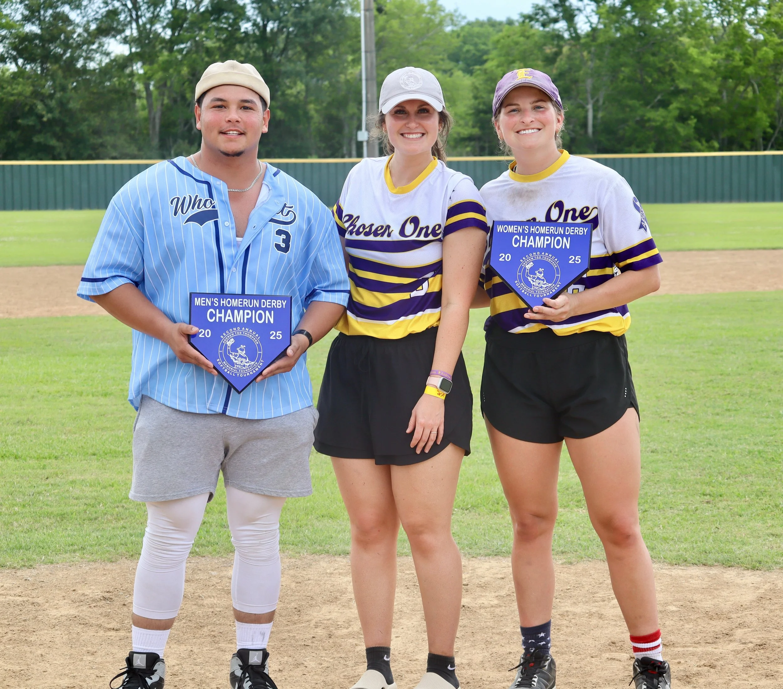 Three young adults standing on a baseball field holding awards for their respective softball and baseball teams, with a green trees background.