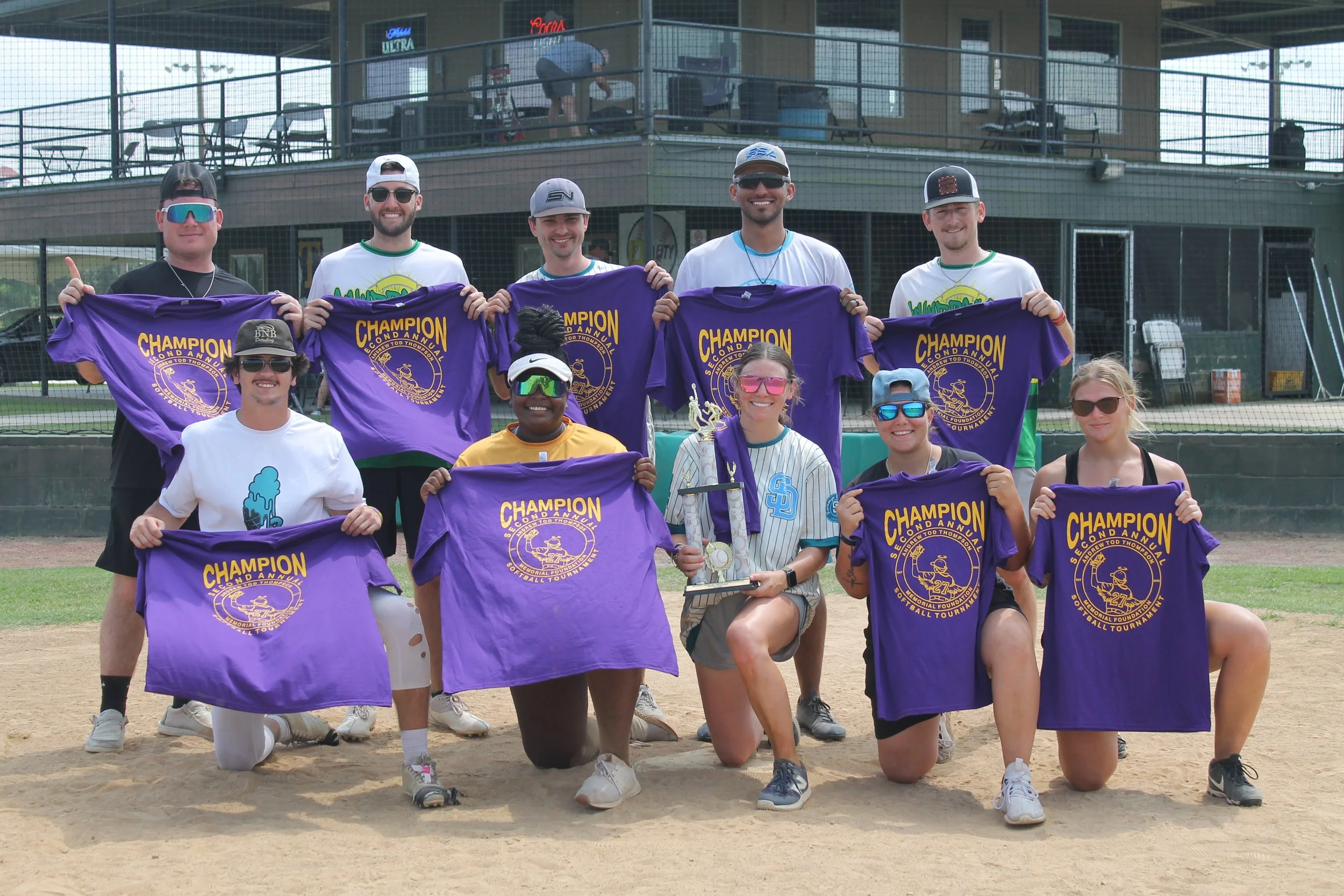 A group of people on a baseball field holding purple t-shirts that say 'Champion' and a logo, posing for a team photo.